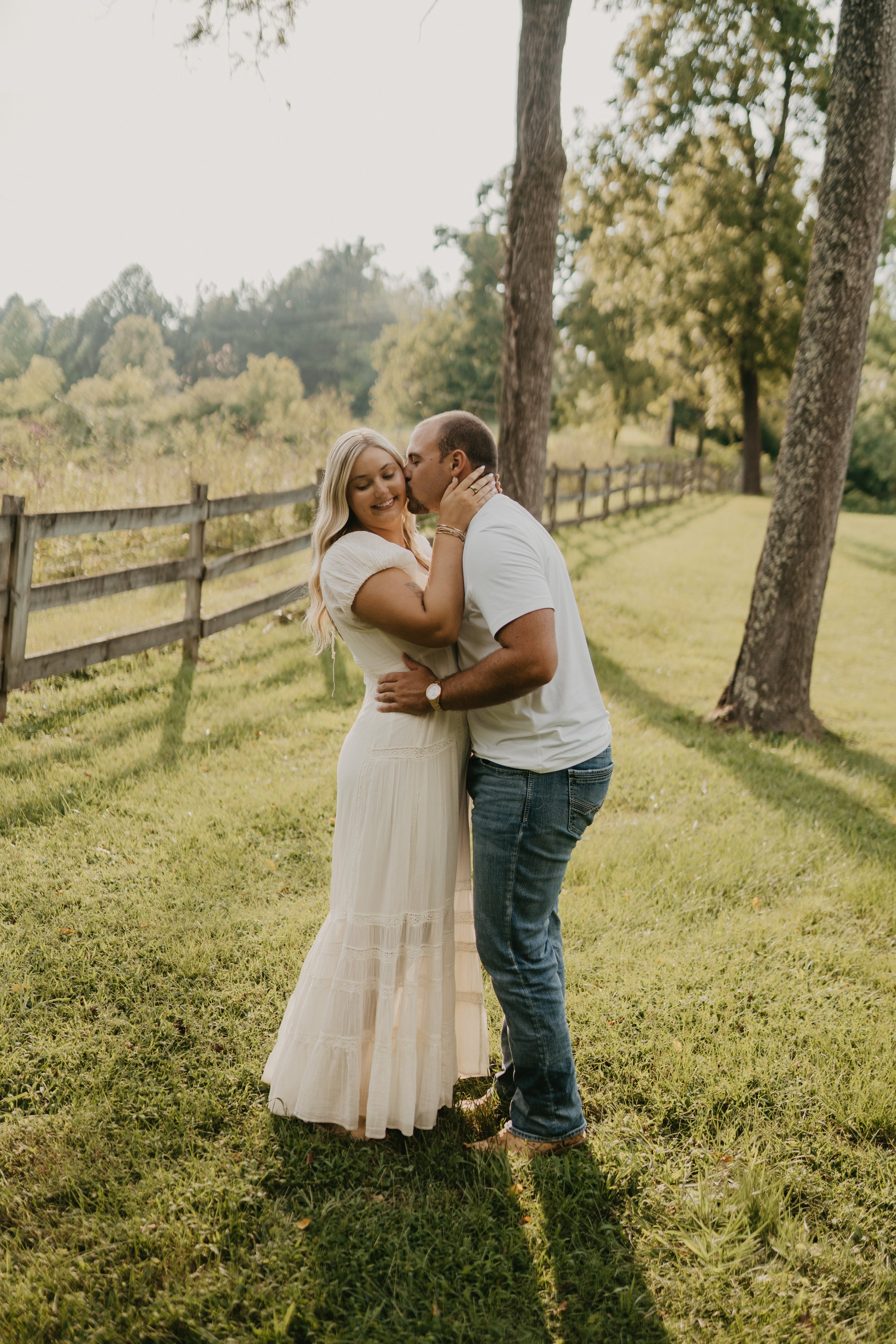 A couple stands outdoors on a grassy area, embracing each other; the woman is smiling with her eyes closed, and the man is kissing her cheek. The woman is wearing a long, cream-colored dress and has long blonde hair, while the man is dressed in a whi