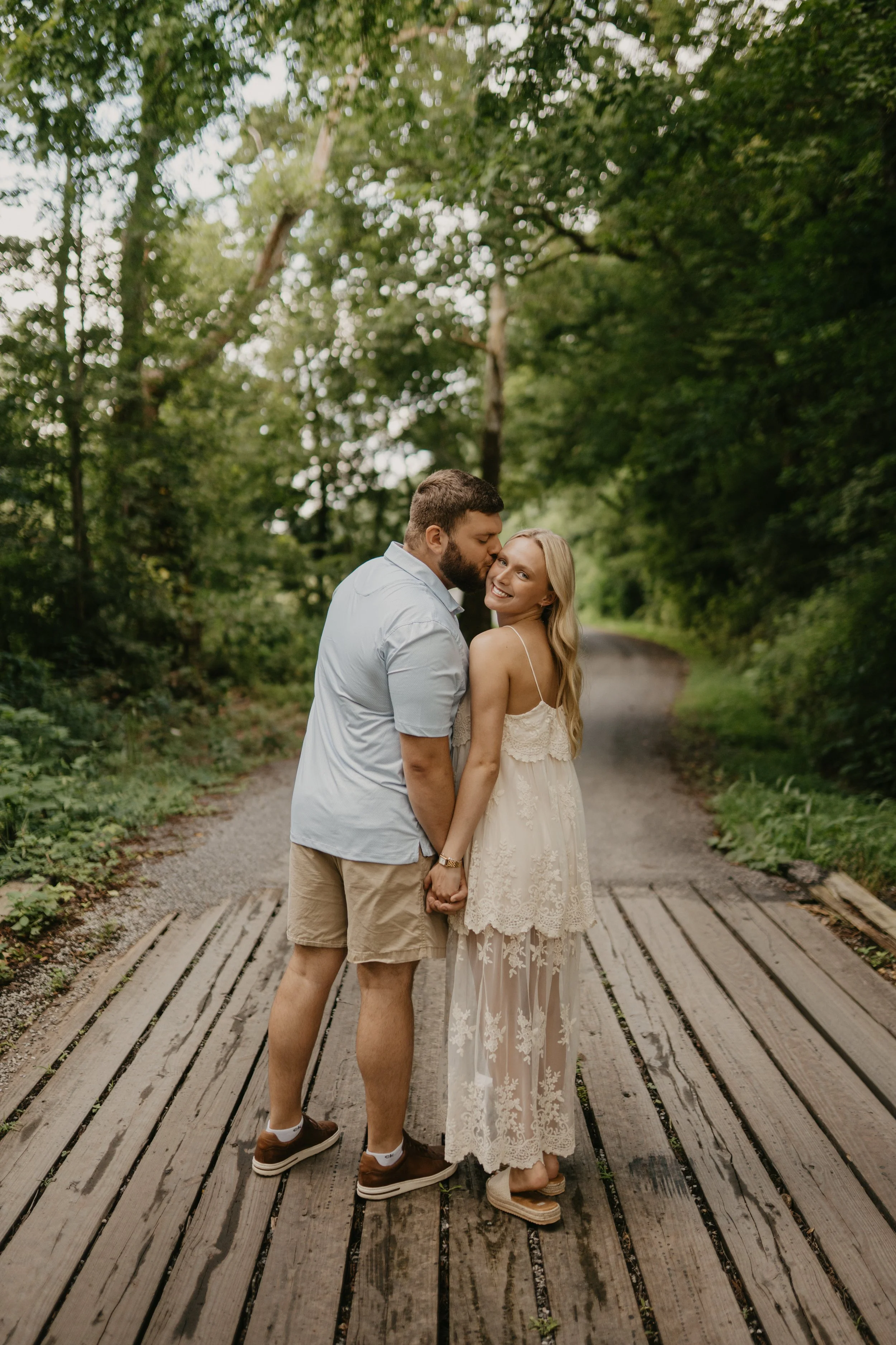 A couple holding hands and sharing a kiss on a wooden bridge in a forested area, with the woman smiling at the camera.