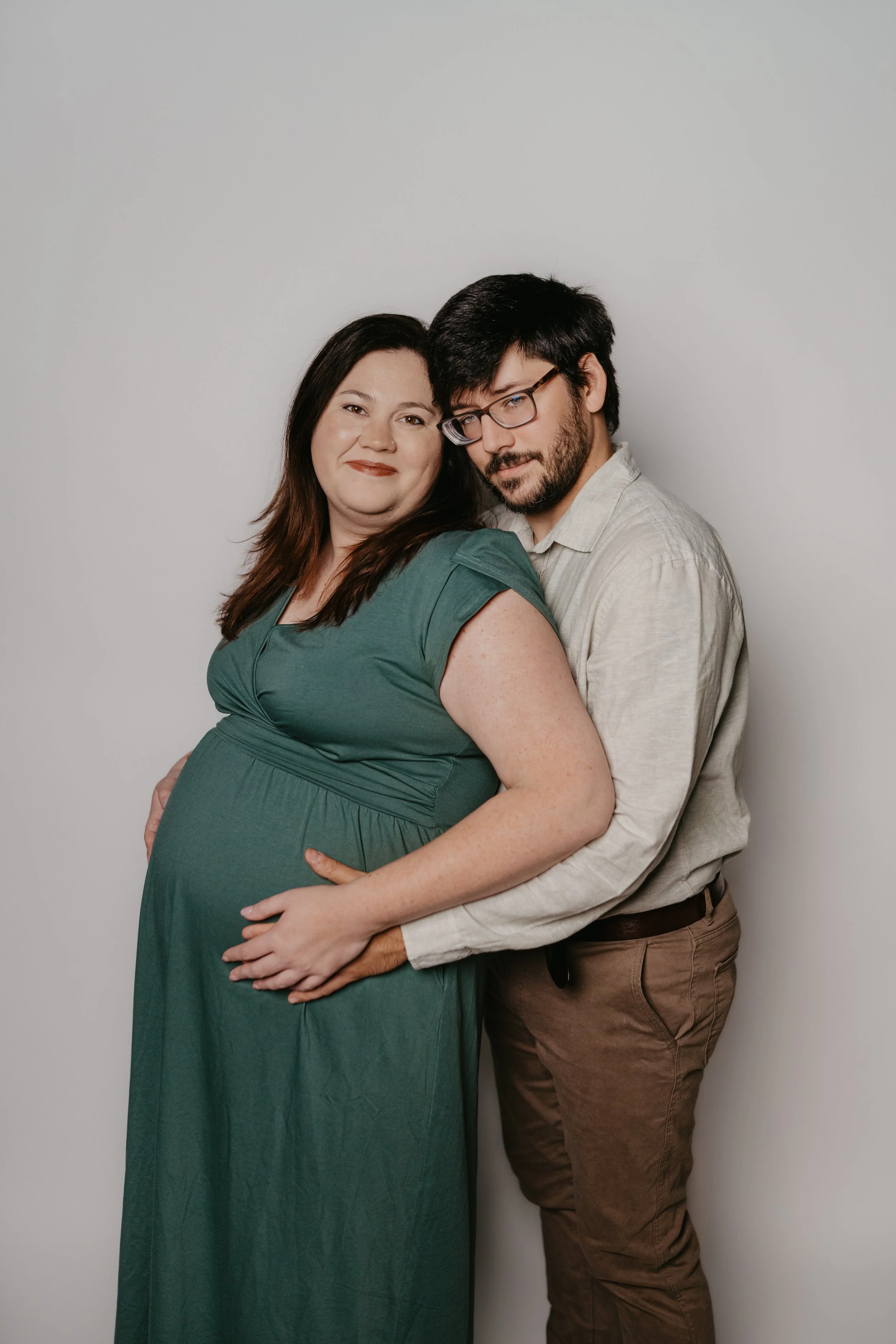 A pregnant woman in a green dress with shoulder-length brown hair smiling, standing beside a man in glasses with dark hair and a beard, wearing a light-colored shirt. He is standing behind her, with his arms around her waist, and both are looking at 