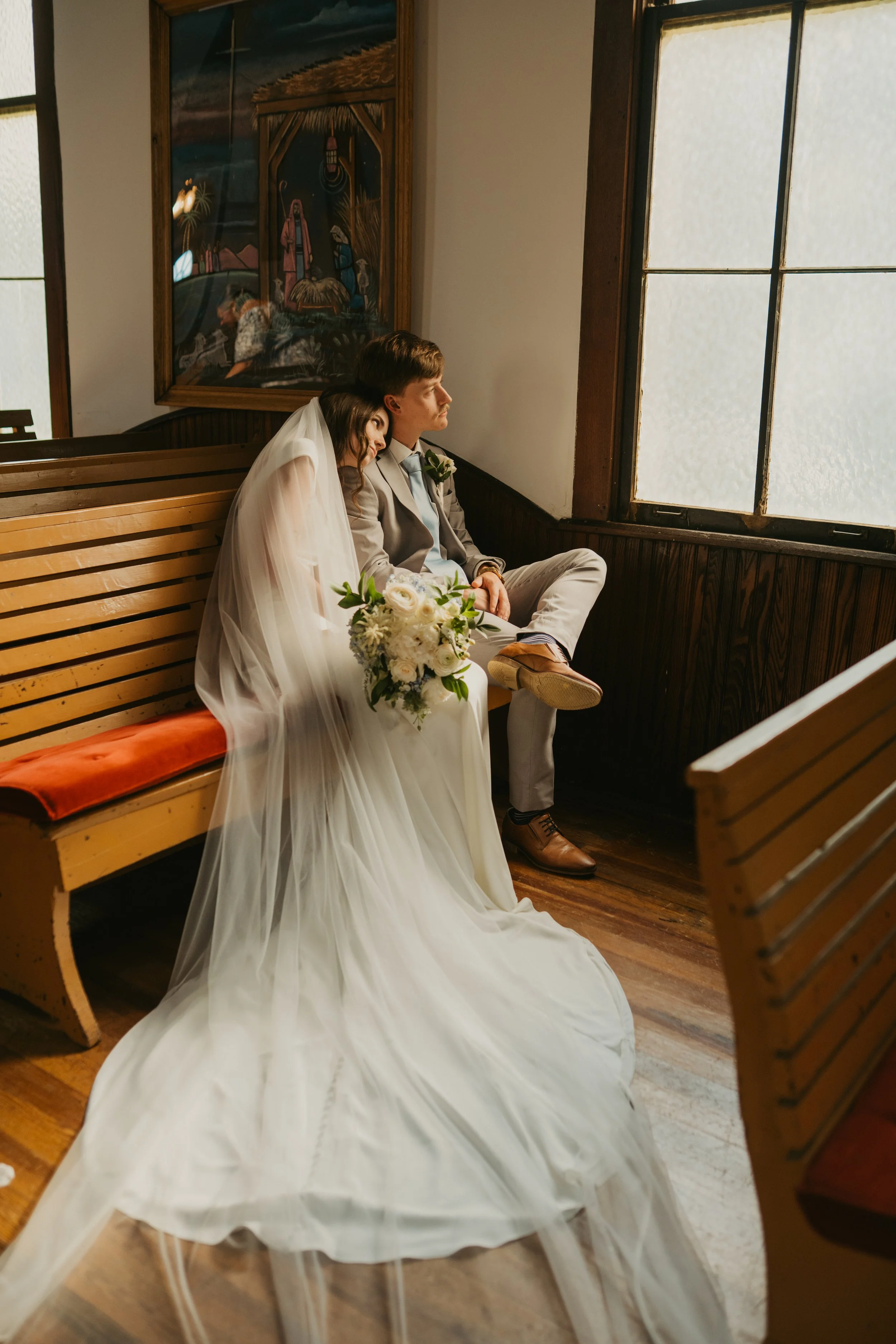 A bride and groom embrace outdoors, with the bride holding a bouquet of pink, white, and yellow flowers. The bride is wearing a lace wedding dress and veil, and the groom is in a gray suit. They are close together, under a clear blue sky with sunlight.