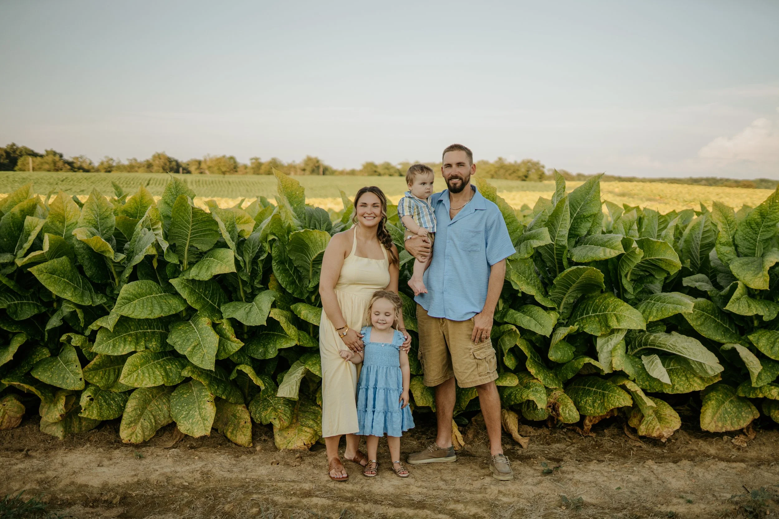 A family of five standing in front of large green tobacco plants in a field with a distant horizon and partially cloudy sky.