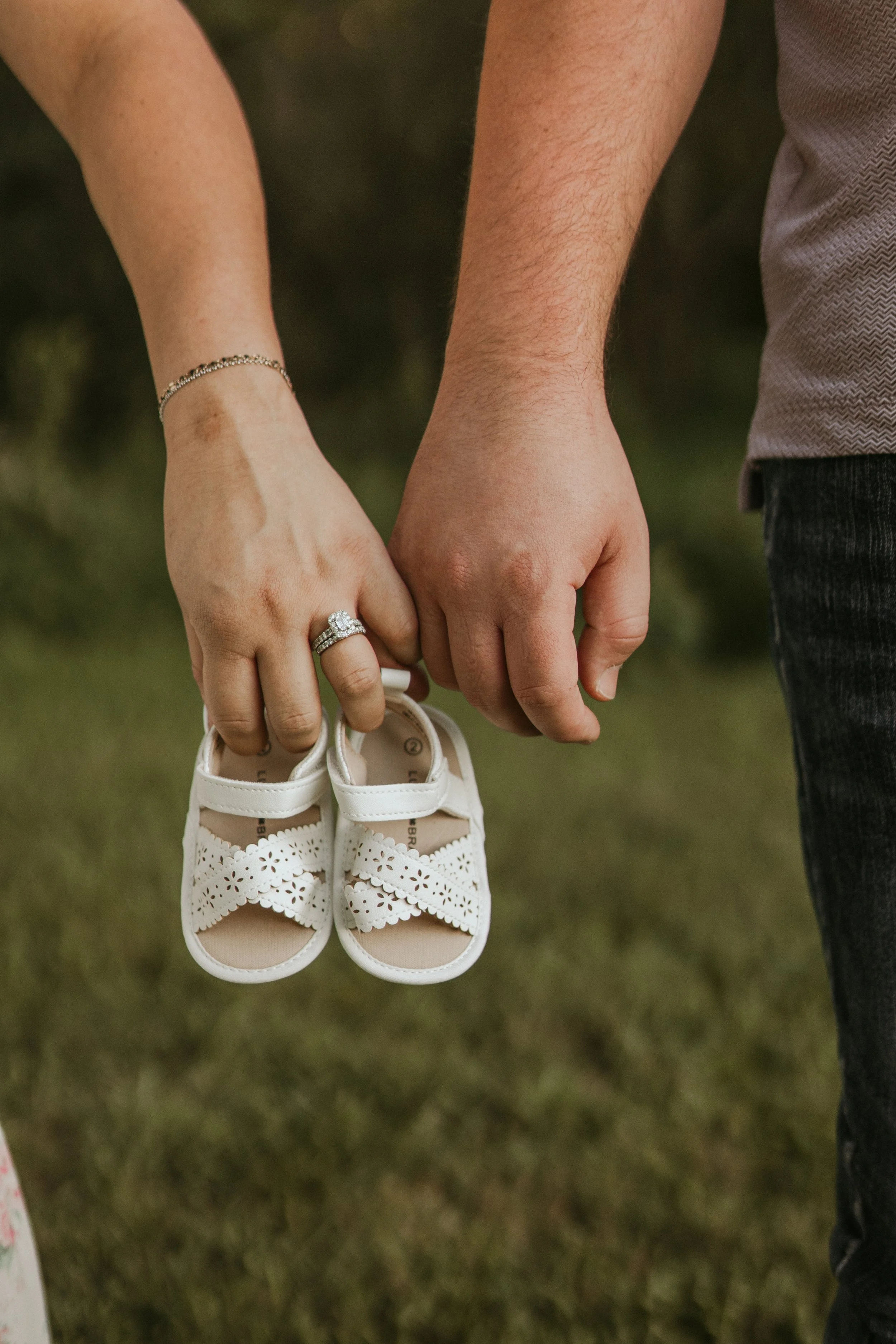 Close-up of a couple holding hands, with the woman's hand hanging downward holding a pair of white baby shoes, and the man's hand holding the woman's hand.