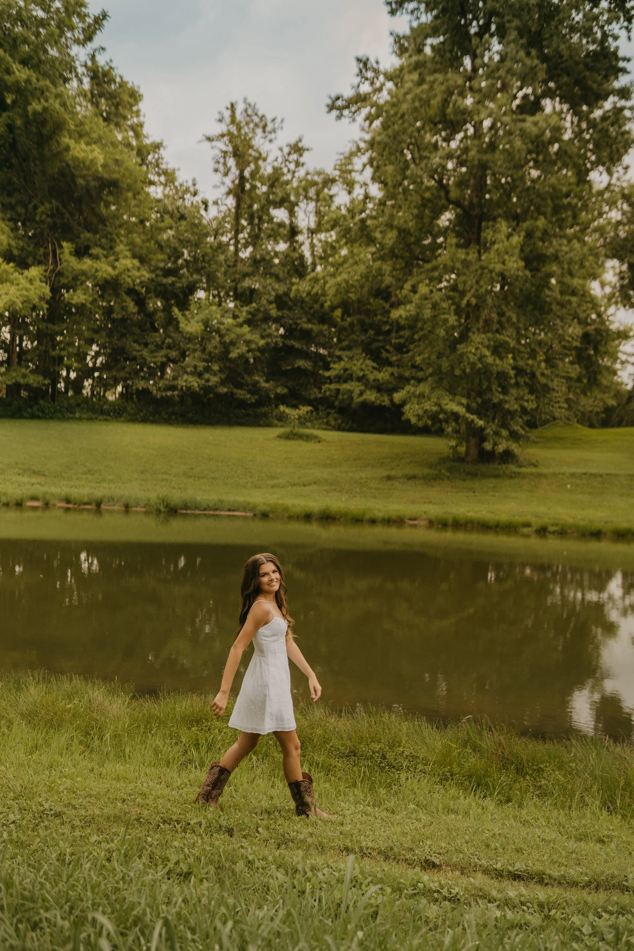 Young woman in white dress walking by a lake with lush green trees in the background during daytime.