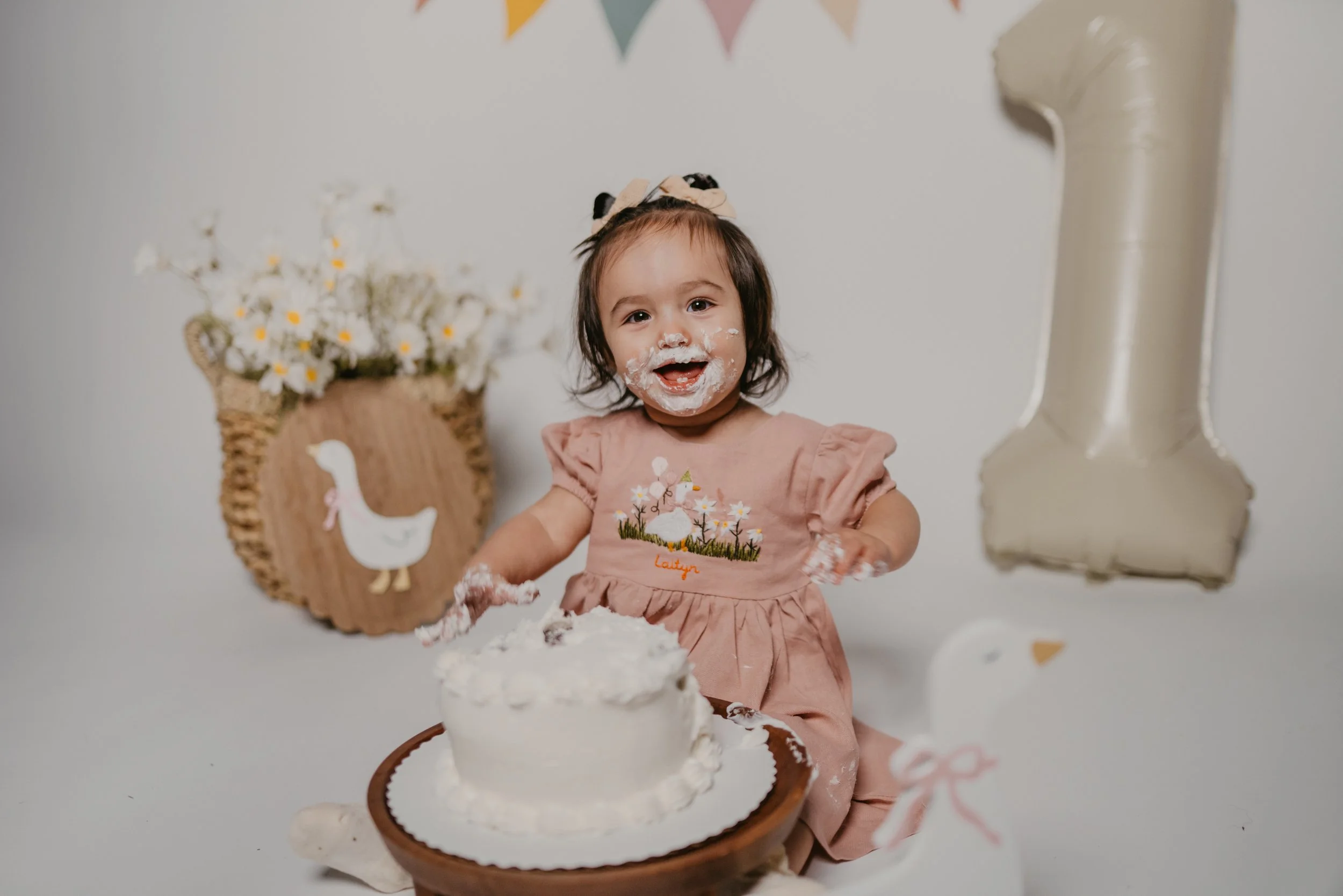 Young girl with brown hair wearing a pink dress with embroidery, celebrating her birthday with a cake with white frosting, decorated with flowers, and a unicorn theme. She has cake and frosting on her face and hands, smiling happily.