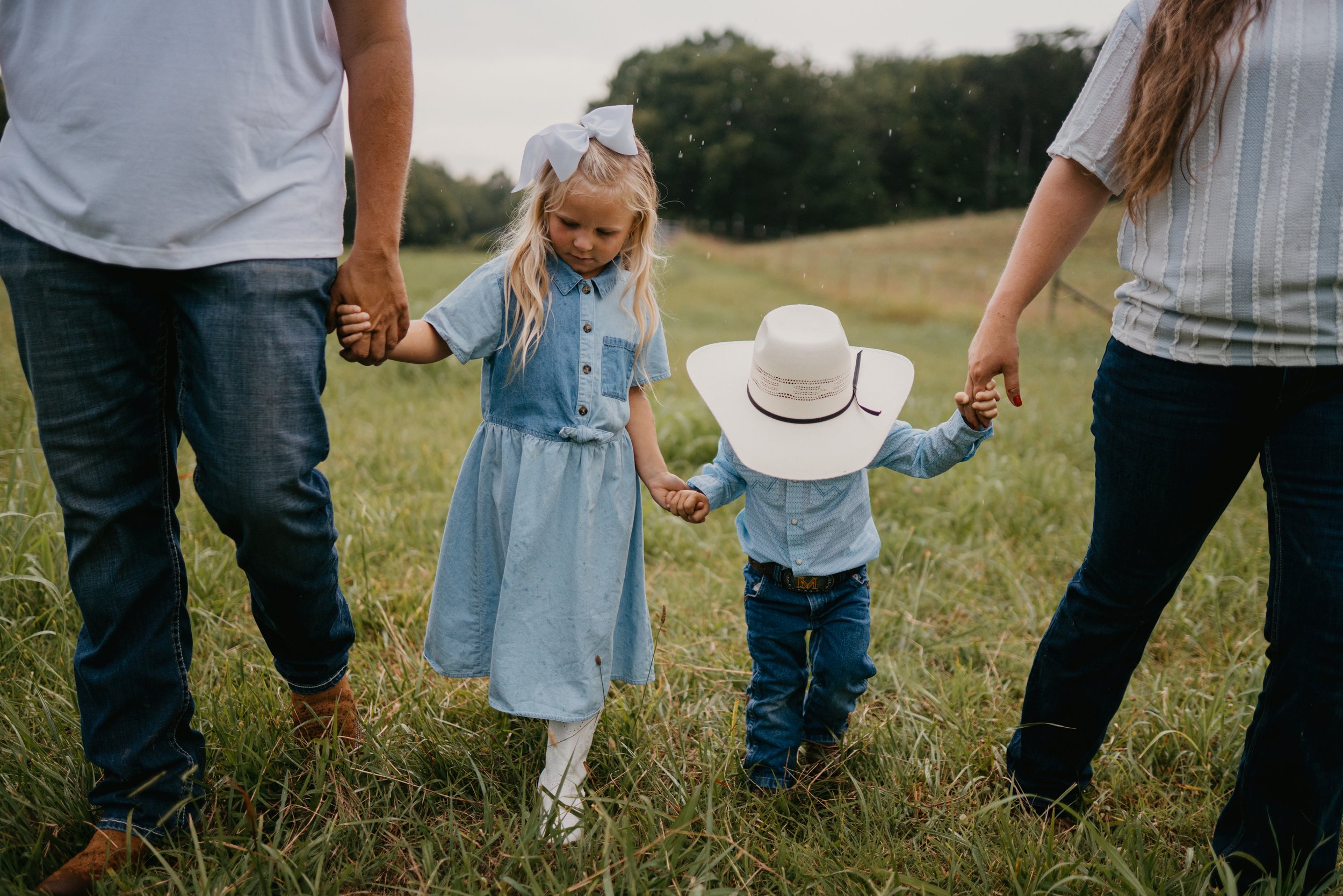 A family holding hands in a grassy field, including two young children with a girl in a blue dress and white bow, and a boy with a white cowboy hat, surrounded by adults, possibly parents, on a cloudy day.