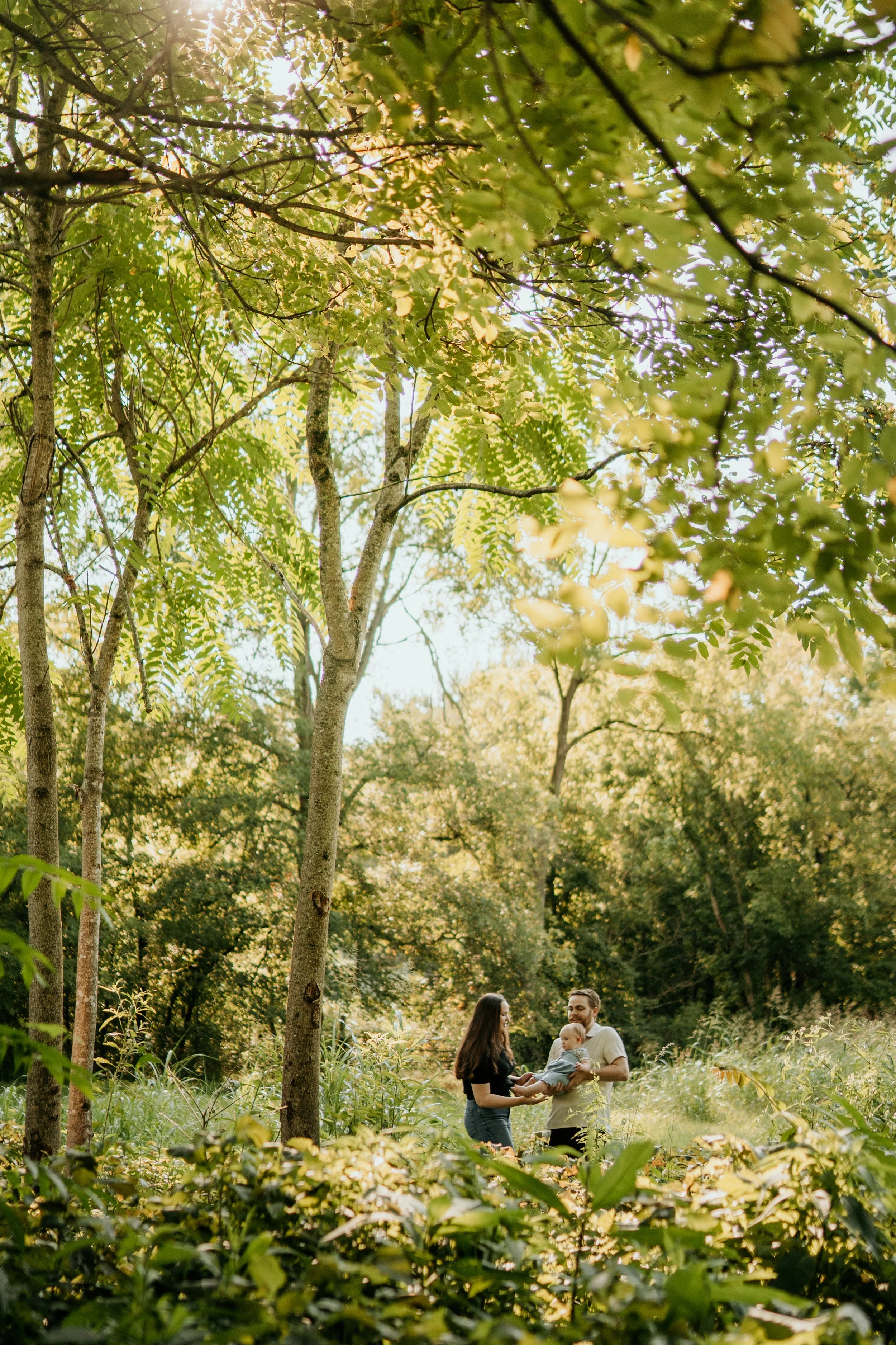 A family of three, including a man, woman, and a baby, standing outdoors in a wooded area with tall trees and green foliage, sharing a moment together.