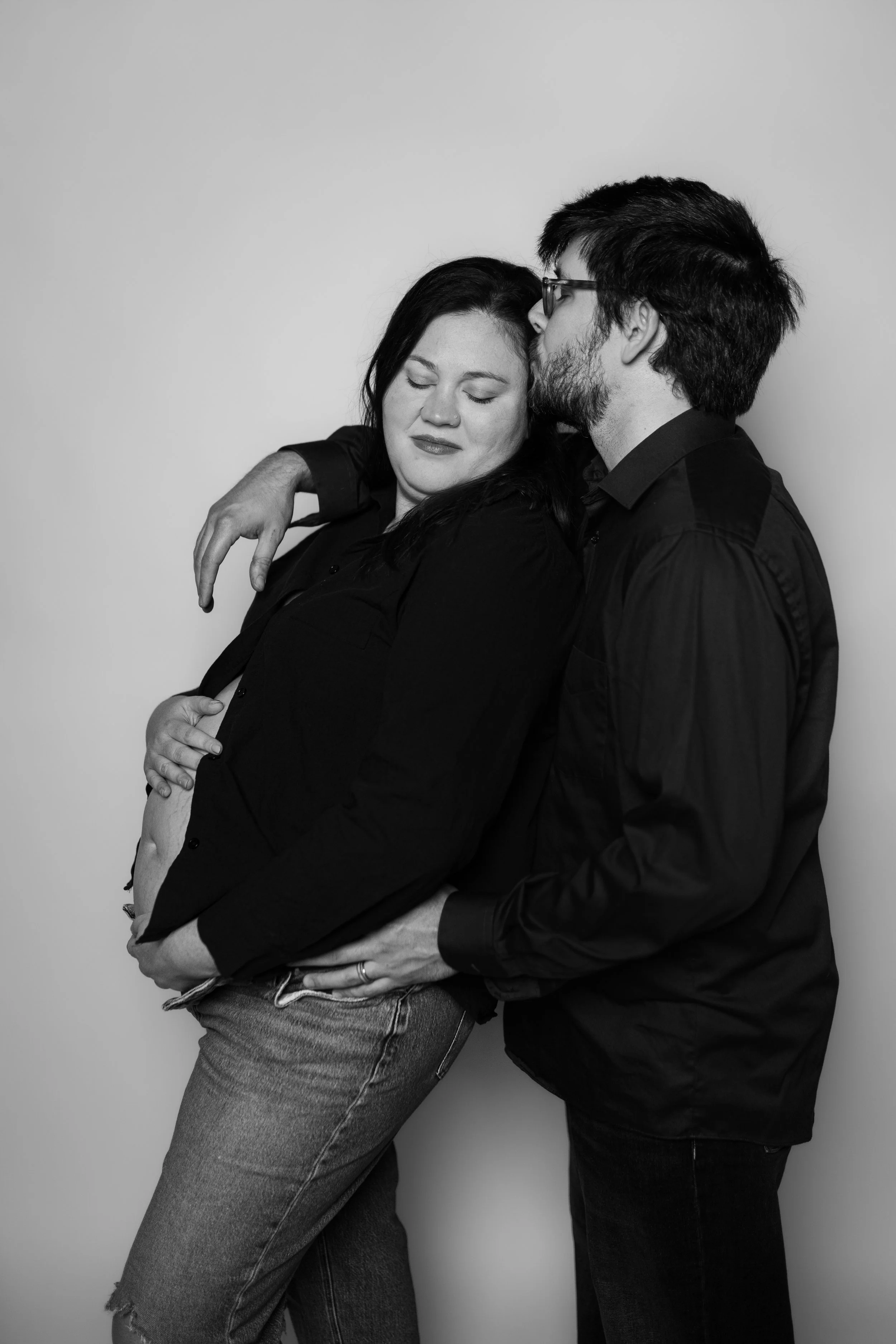Black and white photo of a couple embracing, with the woman closing her eyes and the man kissing her forehead, against a plain white background.
