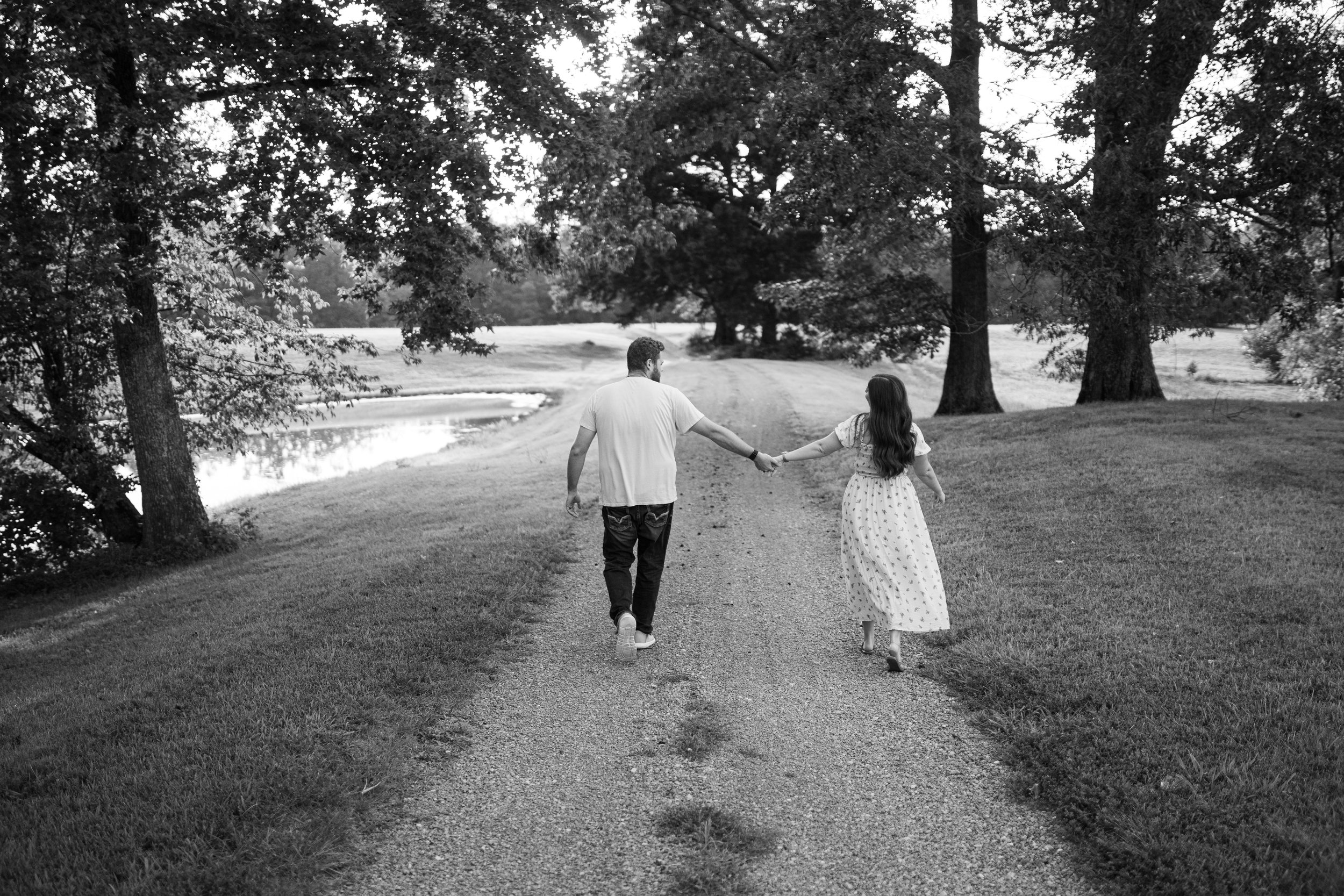 A black-and-white photo of a man and a woman holding hands while walking down a dirt path beside a pond, surrounded by tall trees.