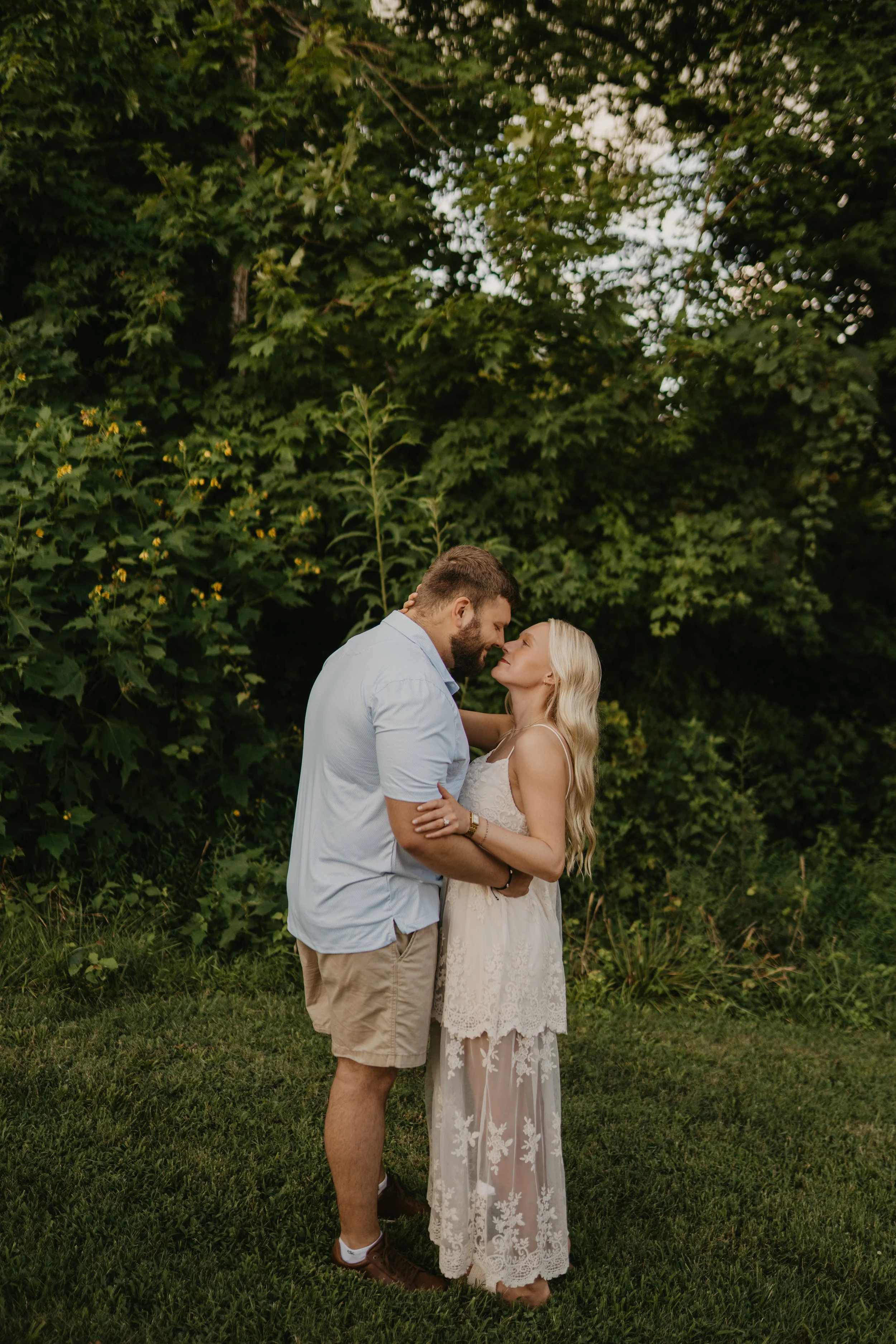 A man and woman stand close together outdoors, gazing into each other's eyes, surrounded by green foliage and grass.