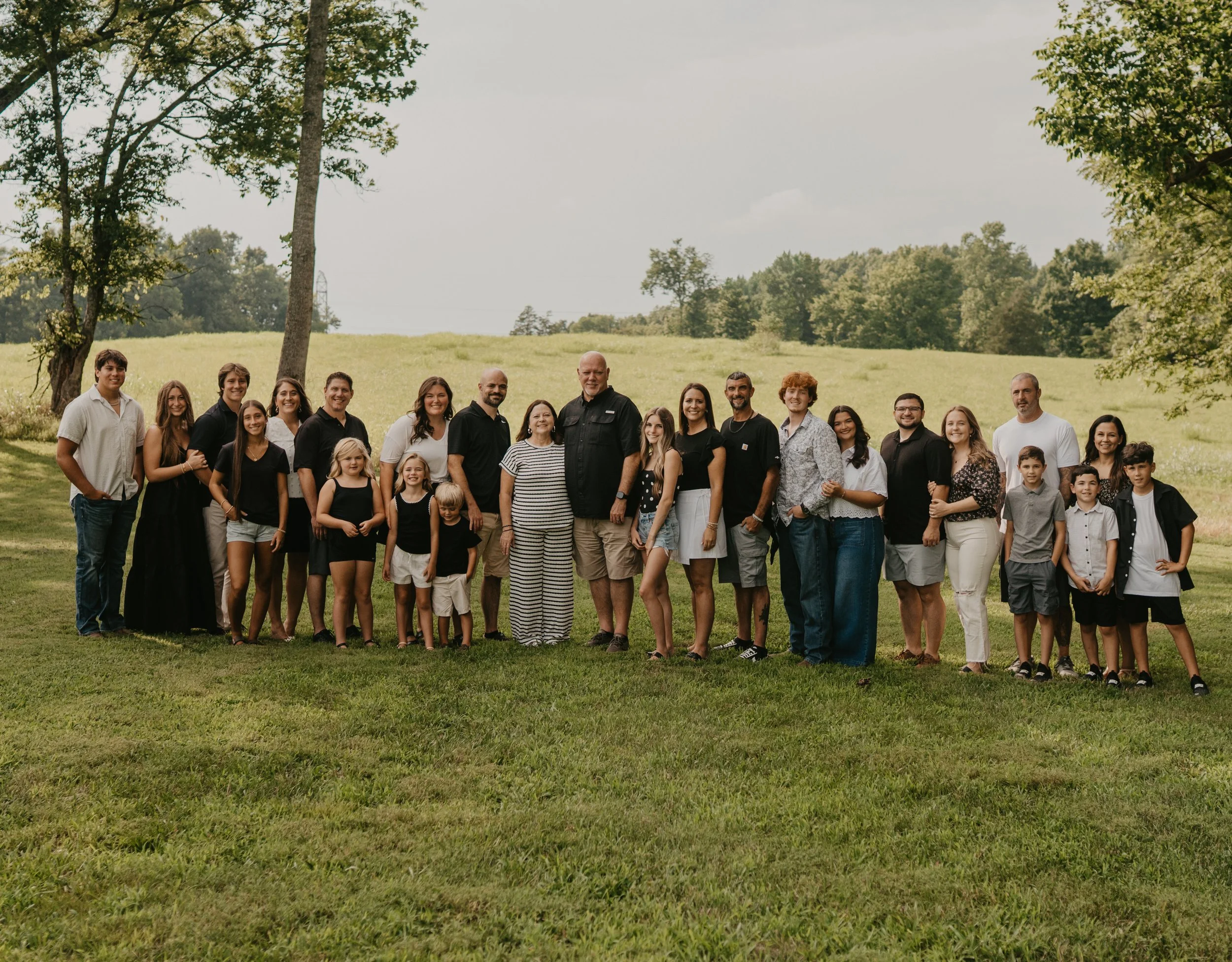 A large group of people including children and adults, standing outdoors on a grassy field with trees and a partly cloudy sky in the background.