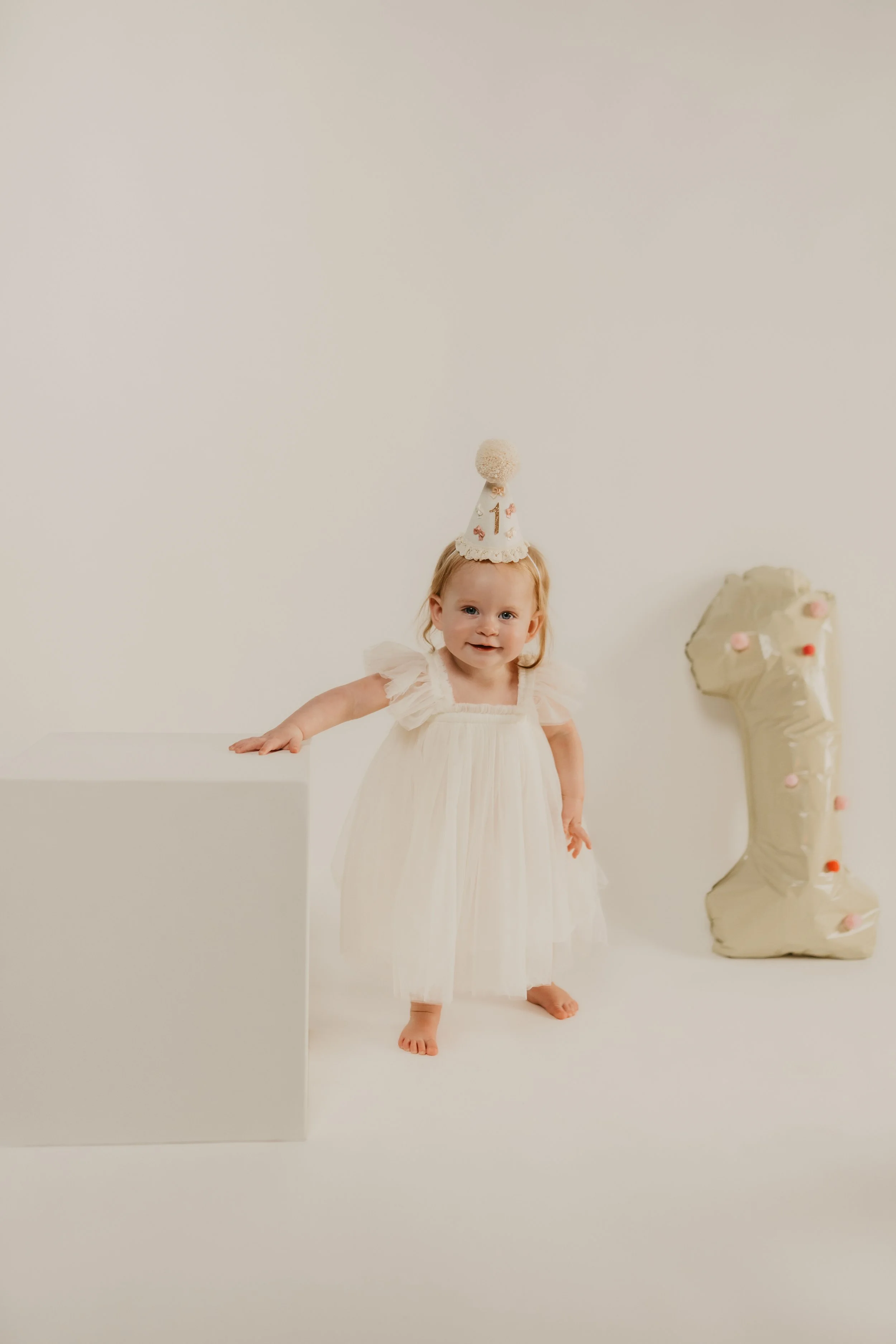 A young girl wearing a white dress and a party hat with a number one on it, standing on a white background with a decorated balloon animal