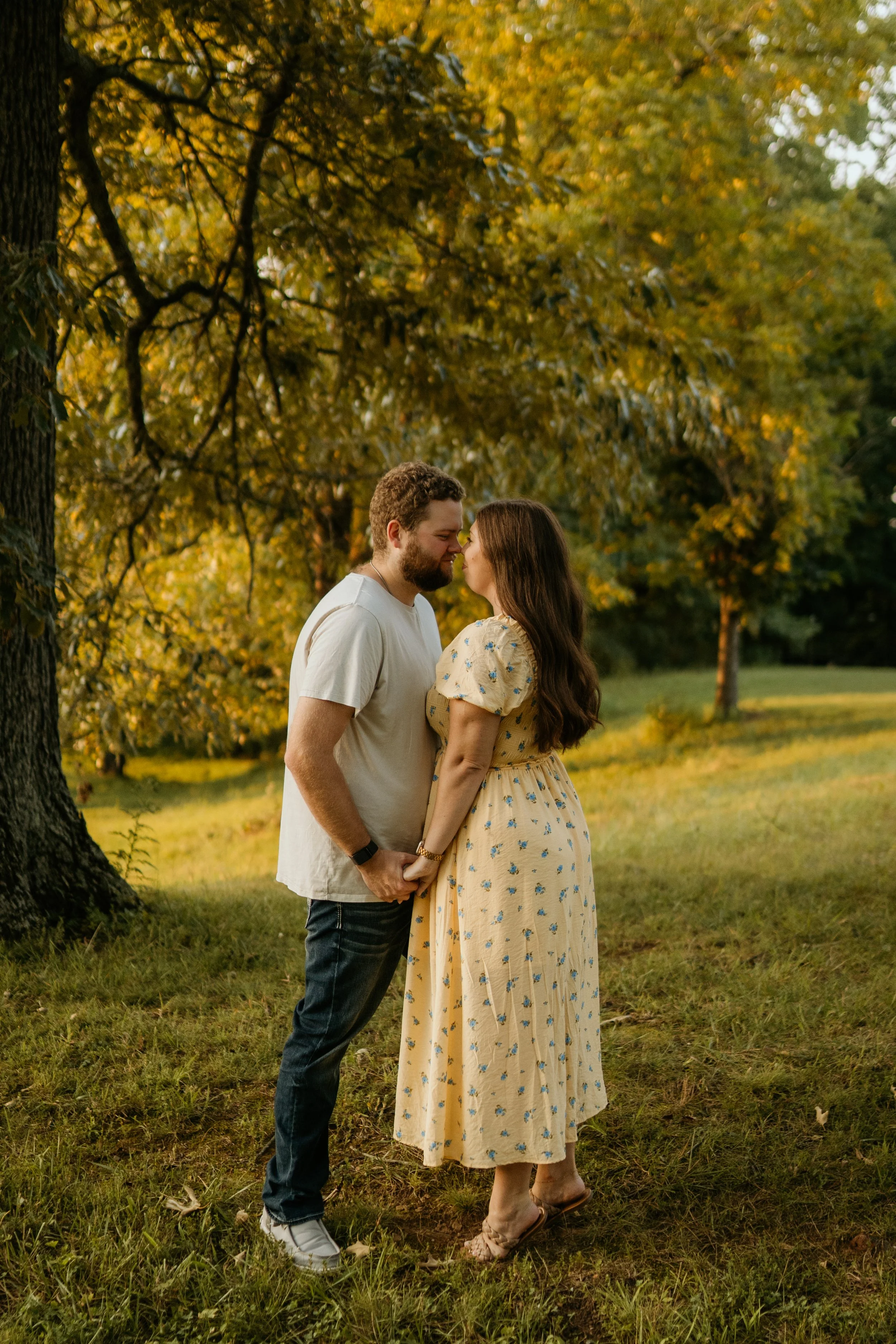 A young couple holding hands and about to kiss outdoors in a park with trees and fall foliage, during golden hour.