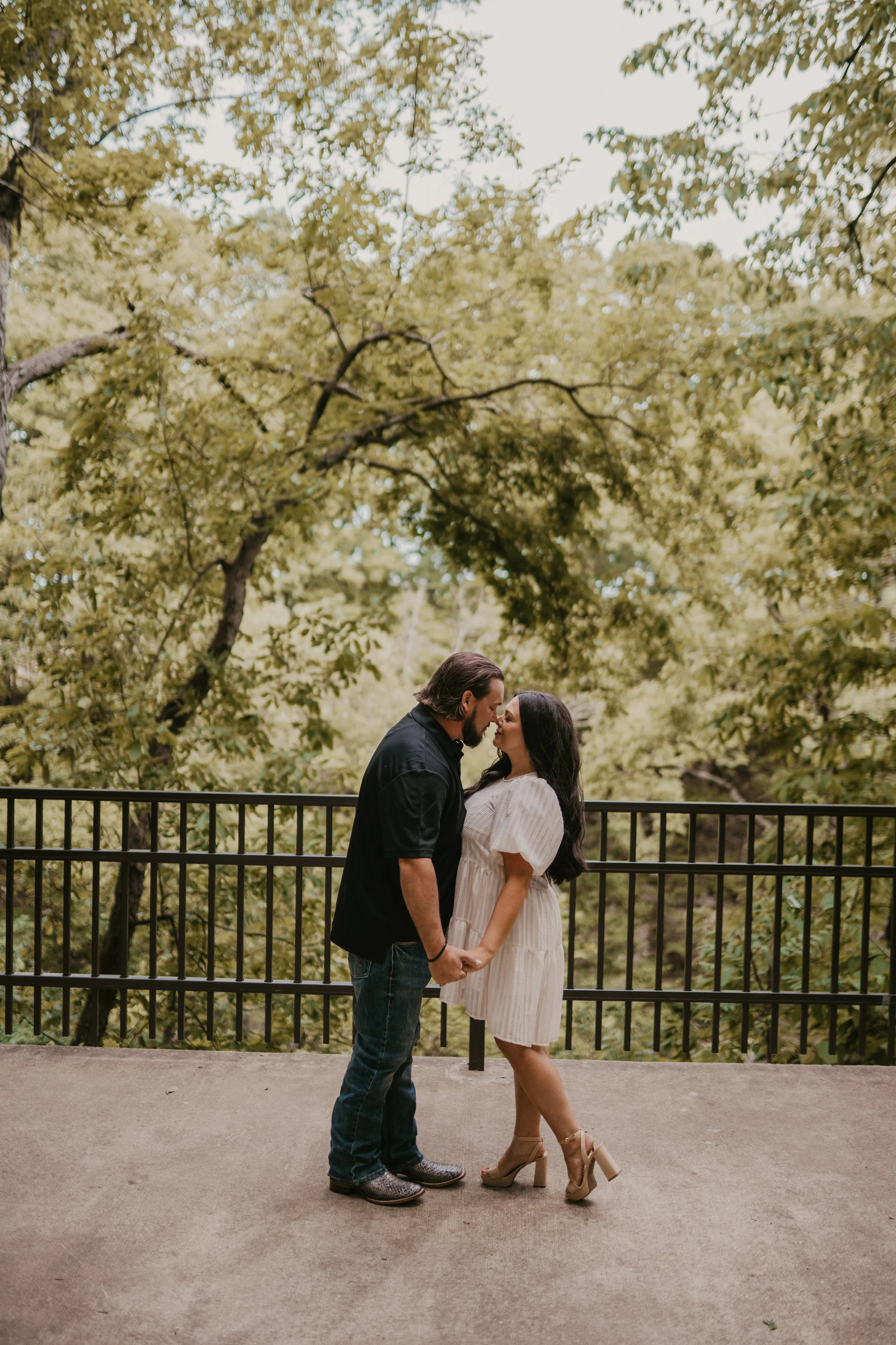 A couple stands close together holding hands, about to kiss on a balcony with a black railing, surrounded by green trees.