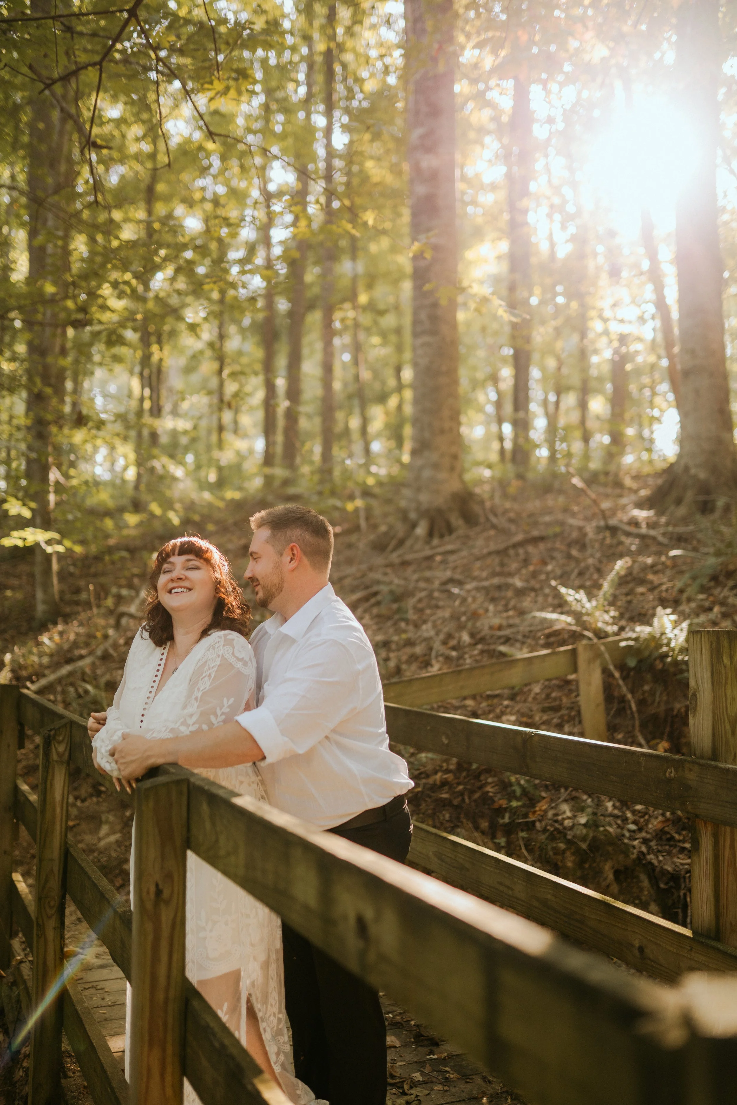 A couple stands on a wooden bridge in a forest, smiling and enjoying the sunlight filtering through the trees.