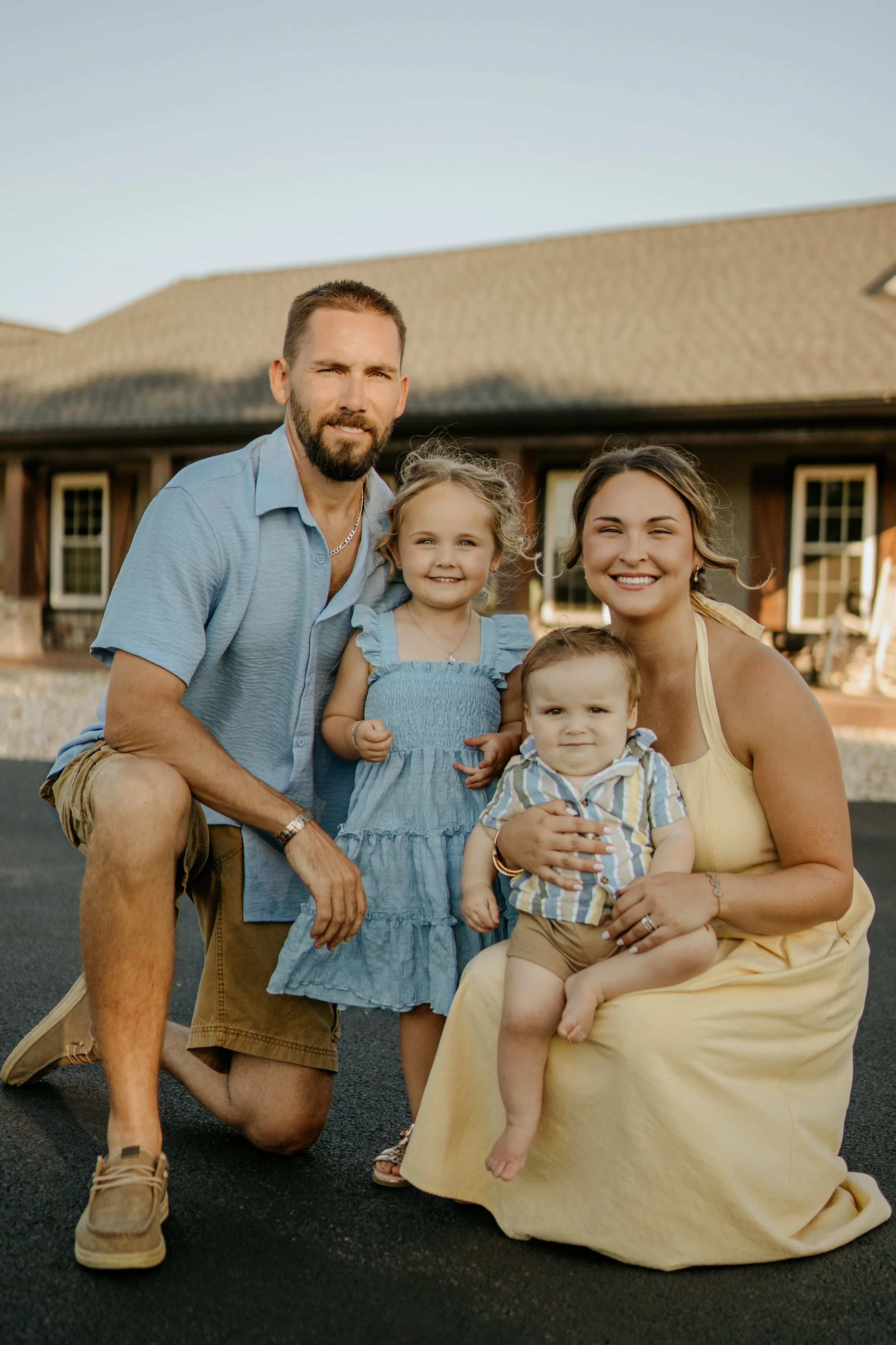 A family of five posing outdoors in front of a house with a brown roof during the daytime. The man with a beard is kneeling on one knee on the left. A woman is sitting on the right, holding a young boy on her lap. Two young girls are standing between
