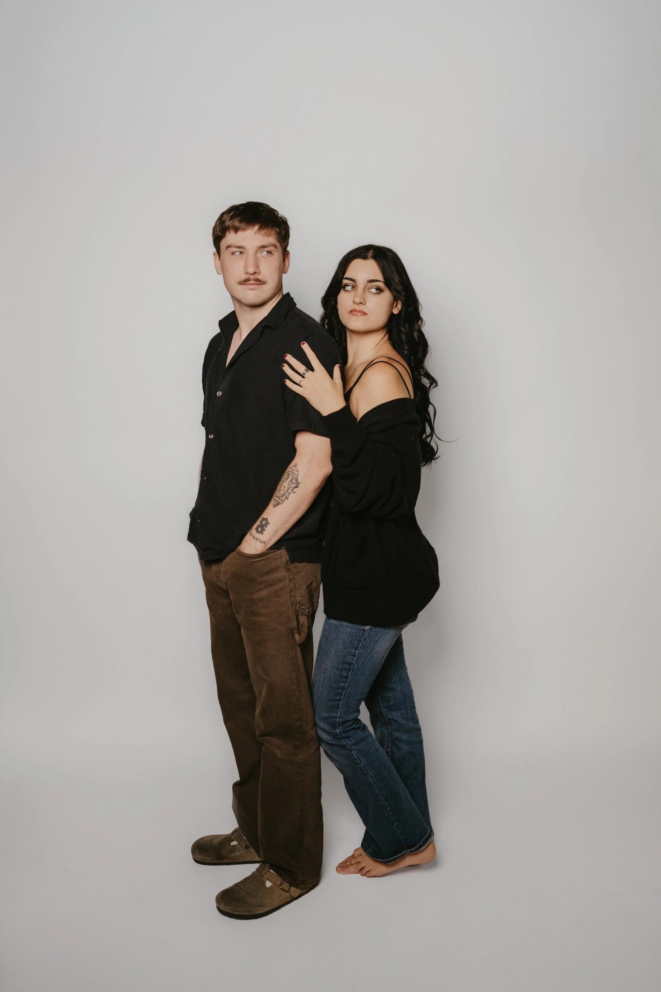A young man and woman are standing close together against a plain light gray background. The man has short brown hair, a mustache, and is dressed in a black shirt and brown pants. The woman has long, dark, wavy hair, and is wearing a black off-the-sh