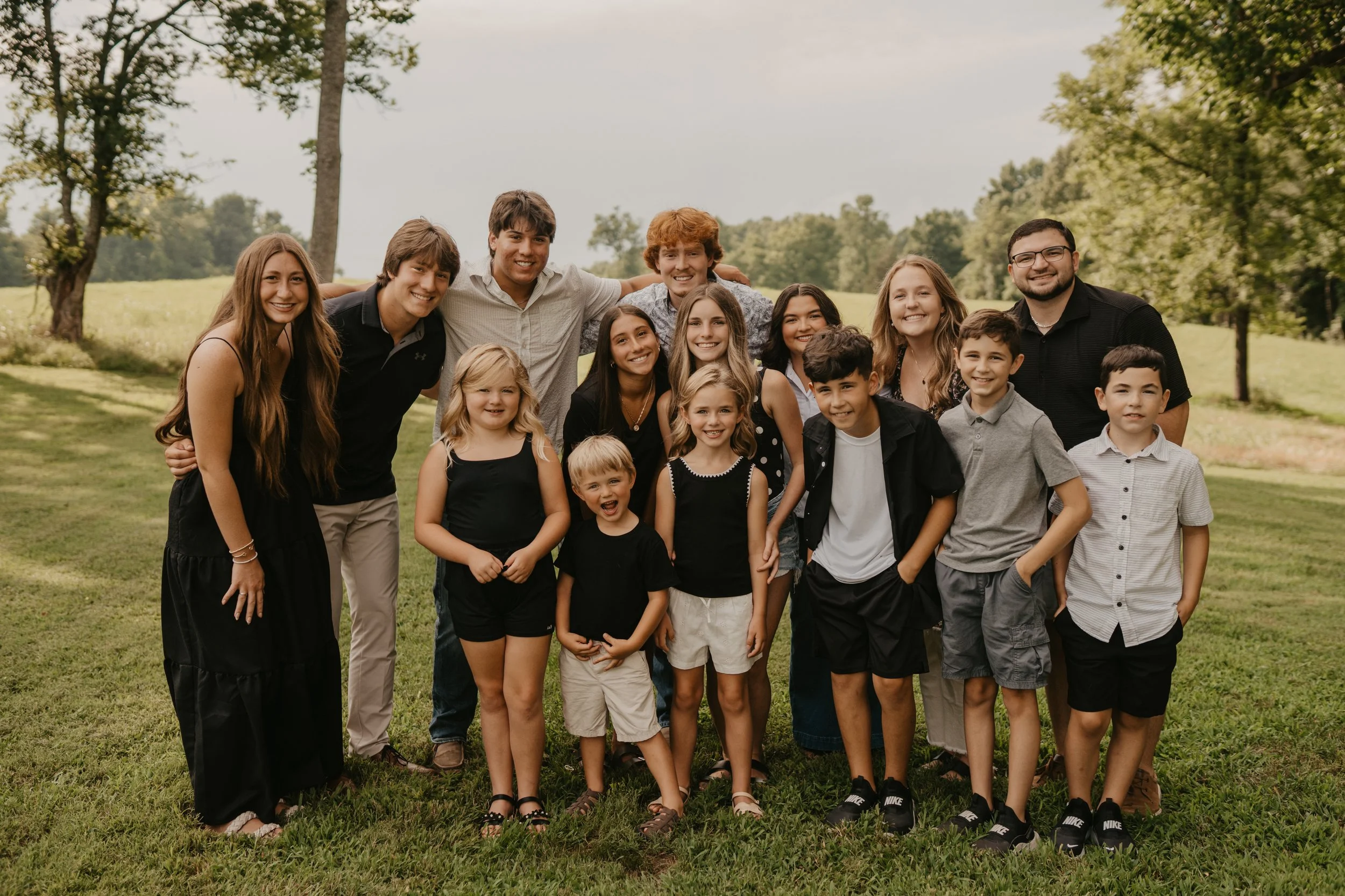 A group of diverse kids and adults smiling outdoors in a park on a sunny day.