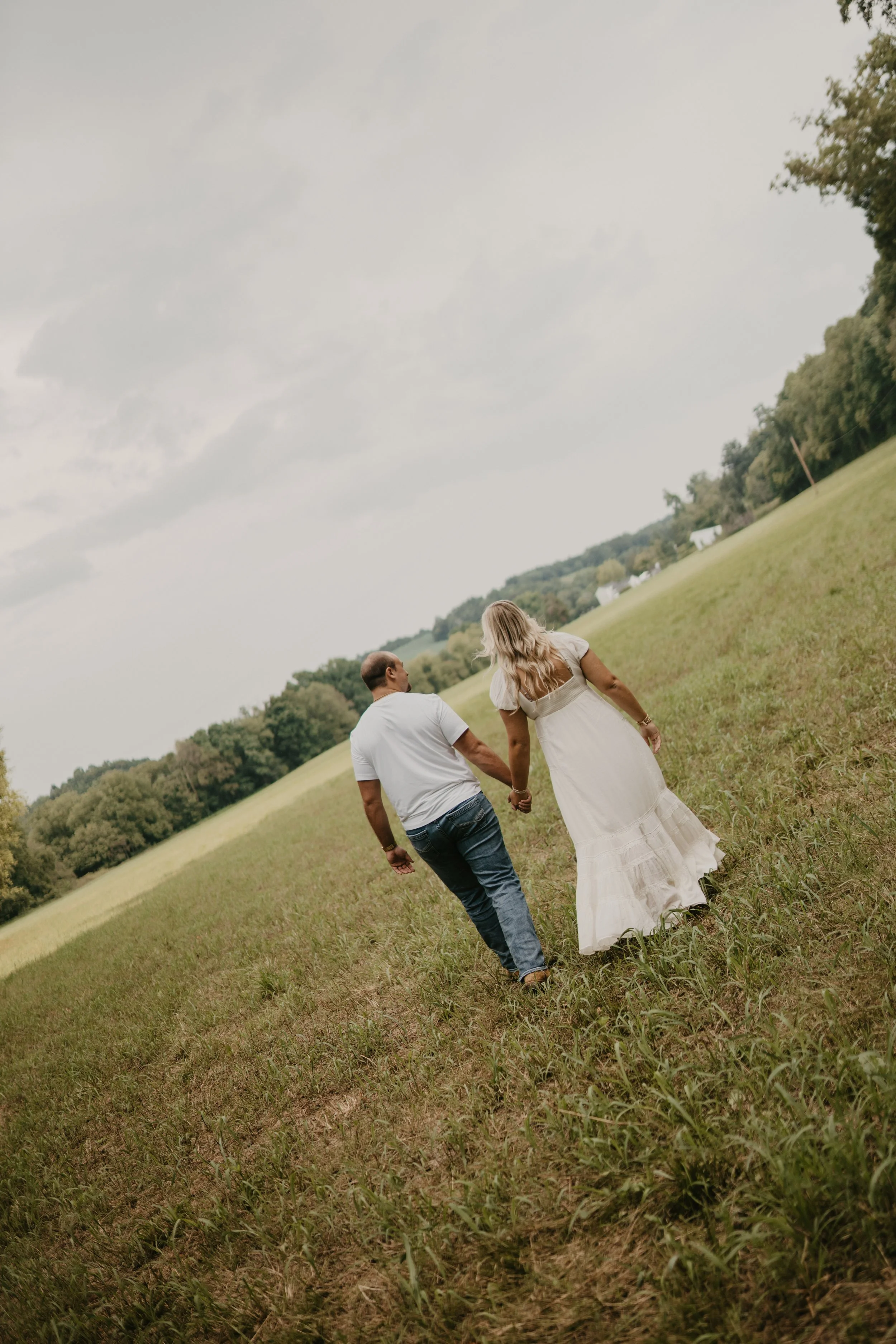 A couple walking hand-in-hand across a grassy field with trees and a cloudy sky in the background.
