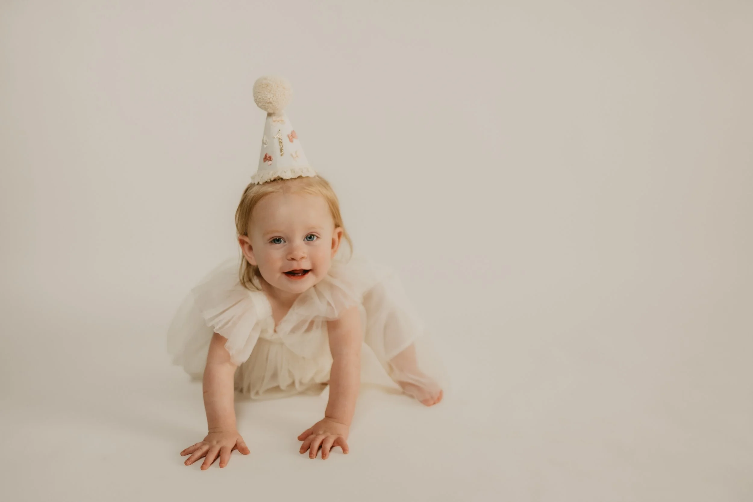 A young girl with blonde hair and blue eyes dressed in a light-colored dress, wearing a polka-dotted party hat with a pom-pom on top, crawling on a plain white background.