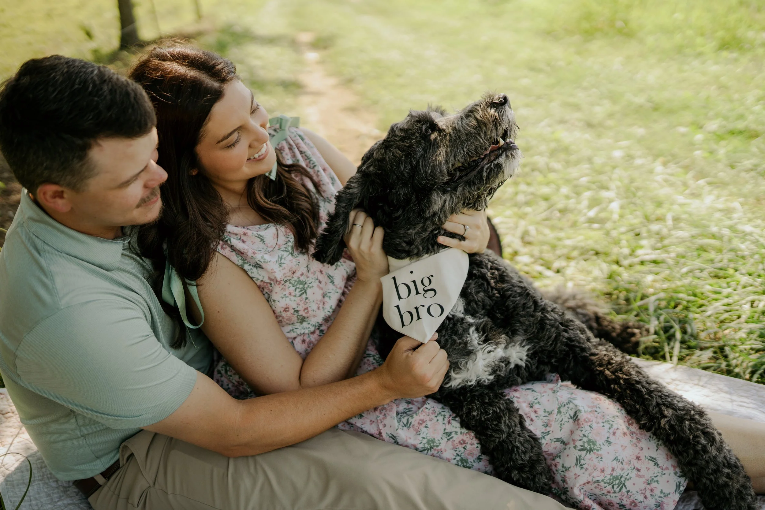 A couple smiling and lying on the grass outdoors with a large, black and gray dog. The dog has a bandana with the text 'big bro' around its neck.