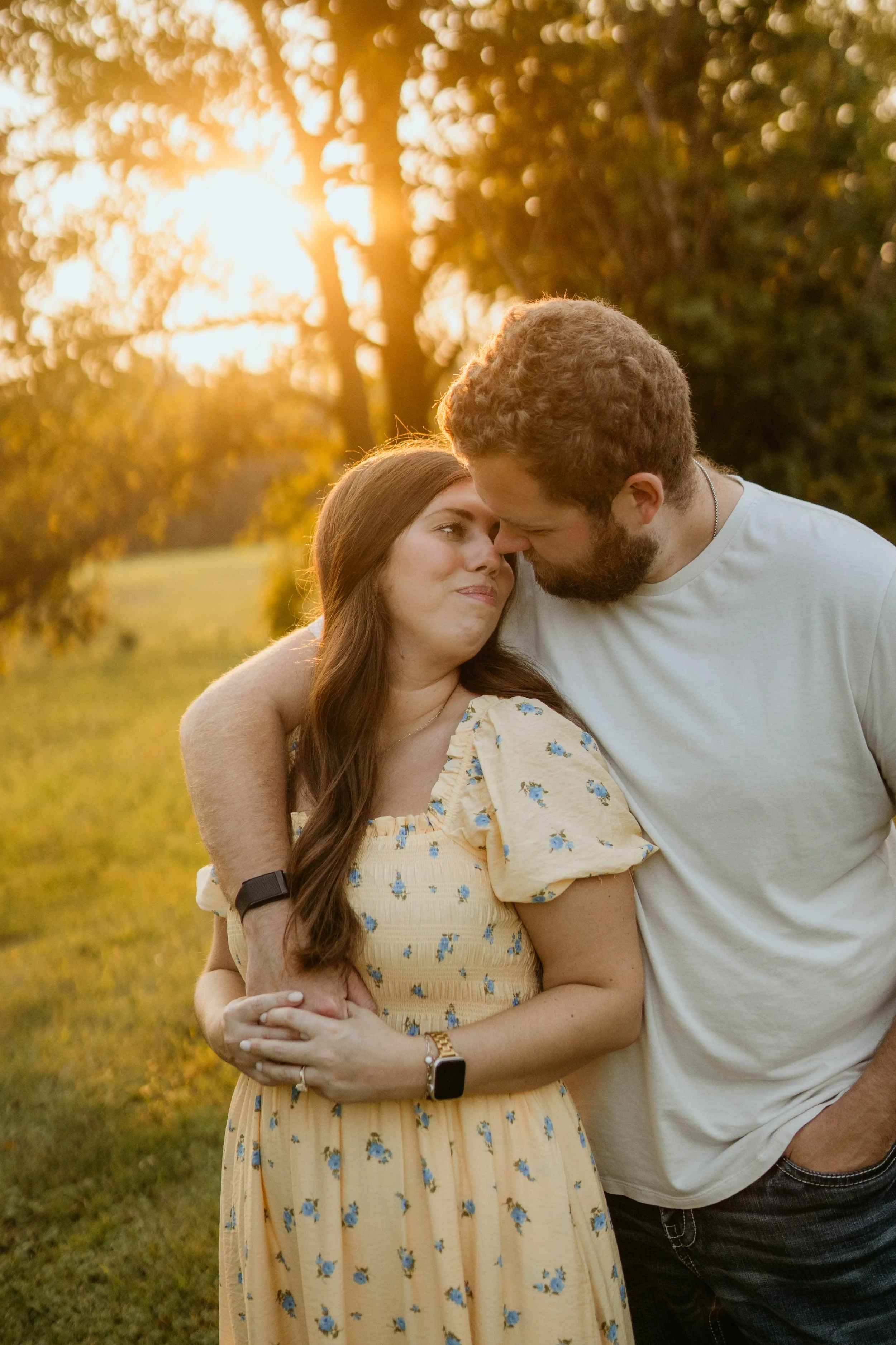 A couple standing close together outdoors during sunset, with the man and woman touching foreheads and sharing a tender moment, surrounded by a natural setting with trees and warm golden light.