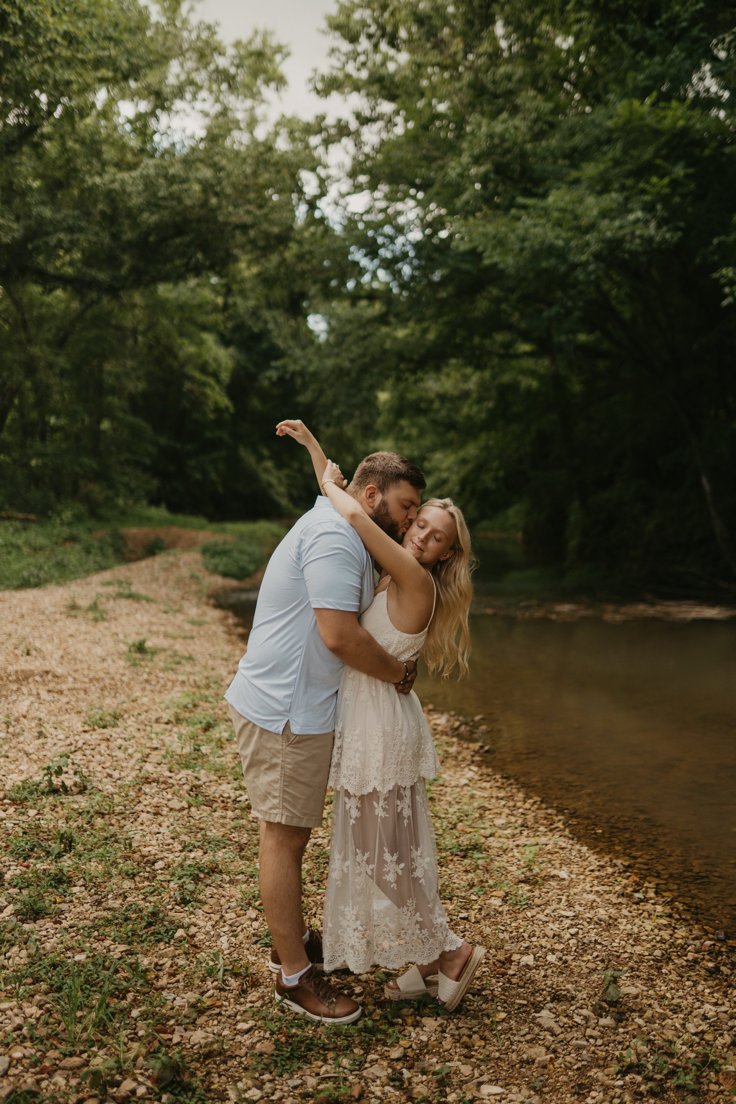 A couple sharing a romantic kiss by a river during daytime, surrounded by trees.