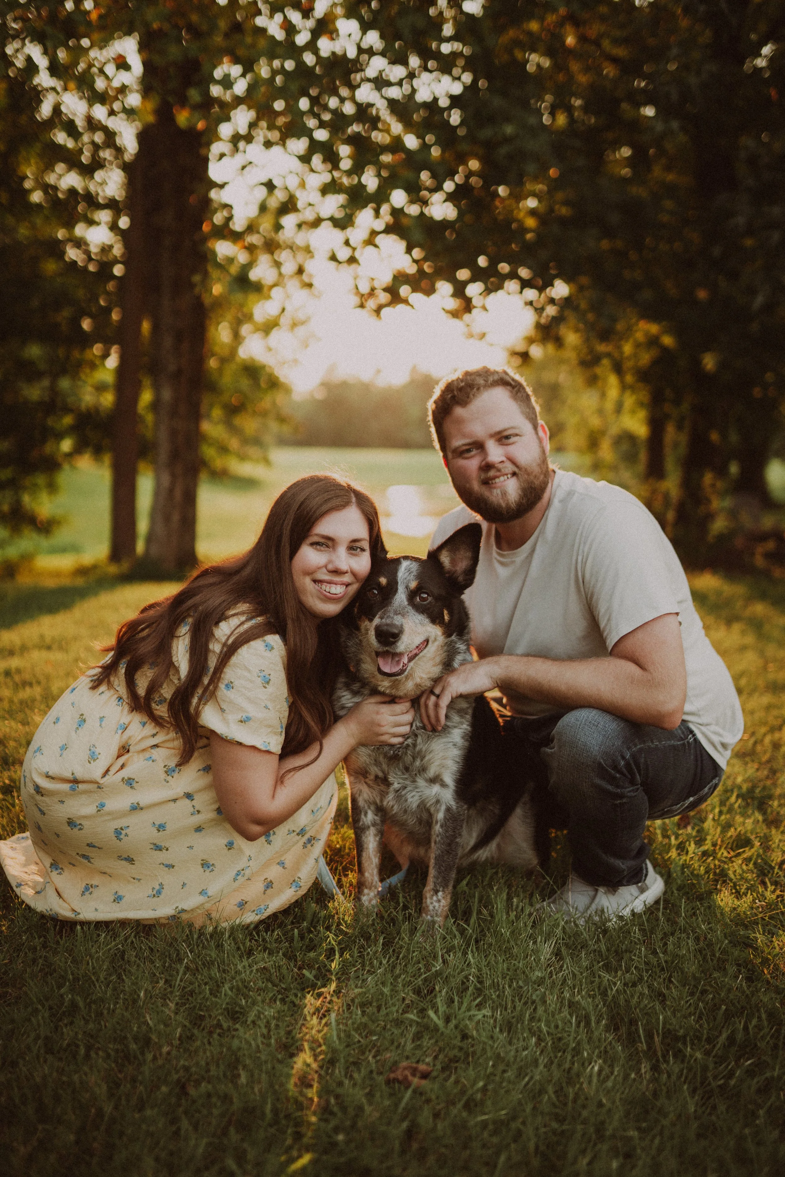 A young woman and a young man smiling while kneeling with their dog in a grassy park during sunset, with trees and warm sunlight in the background.