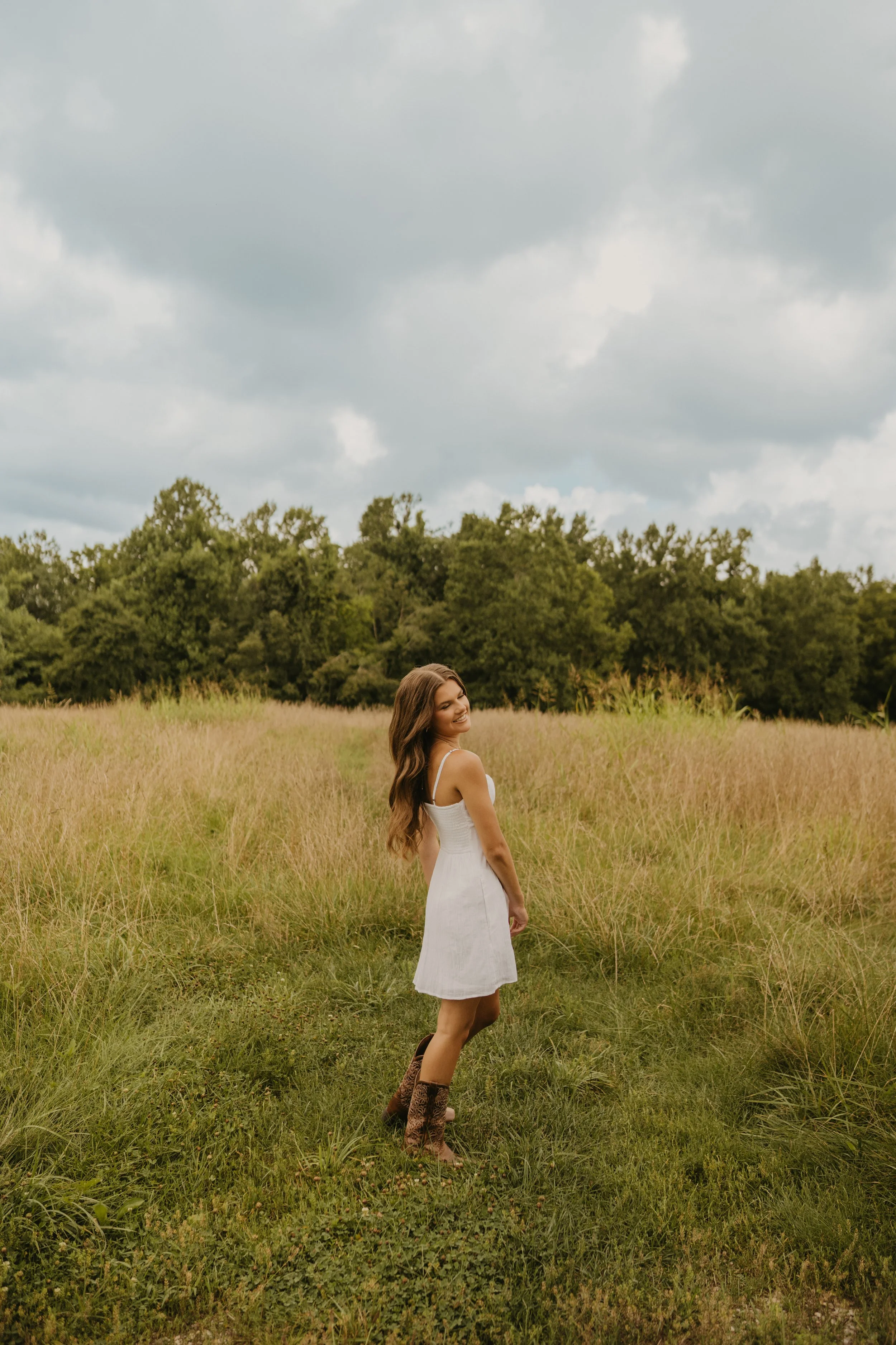 A young woman with long brown hair in a white dress and cowboy boots stands in a grassy field, smiling, with trees and a cloudy sky in the background.