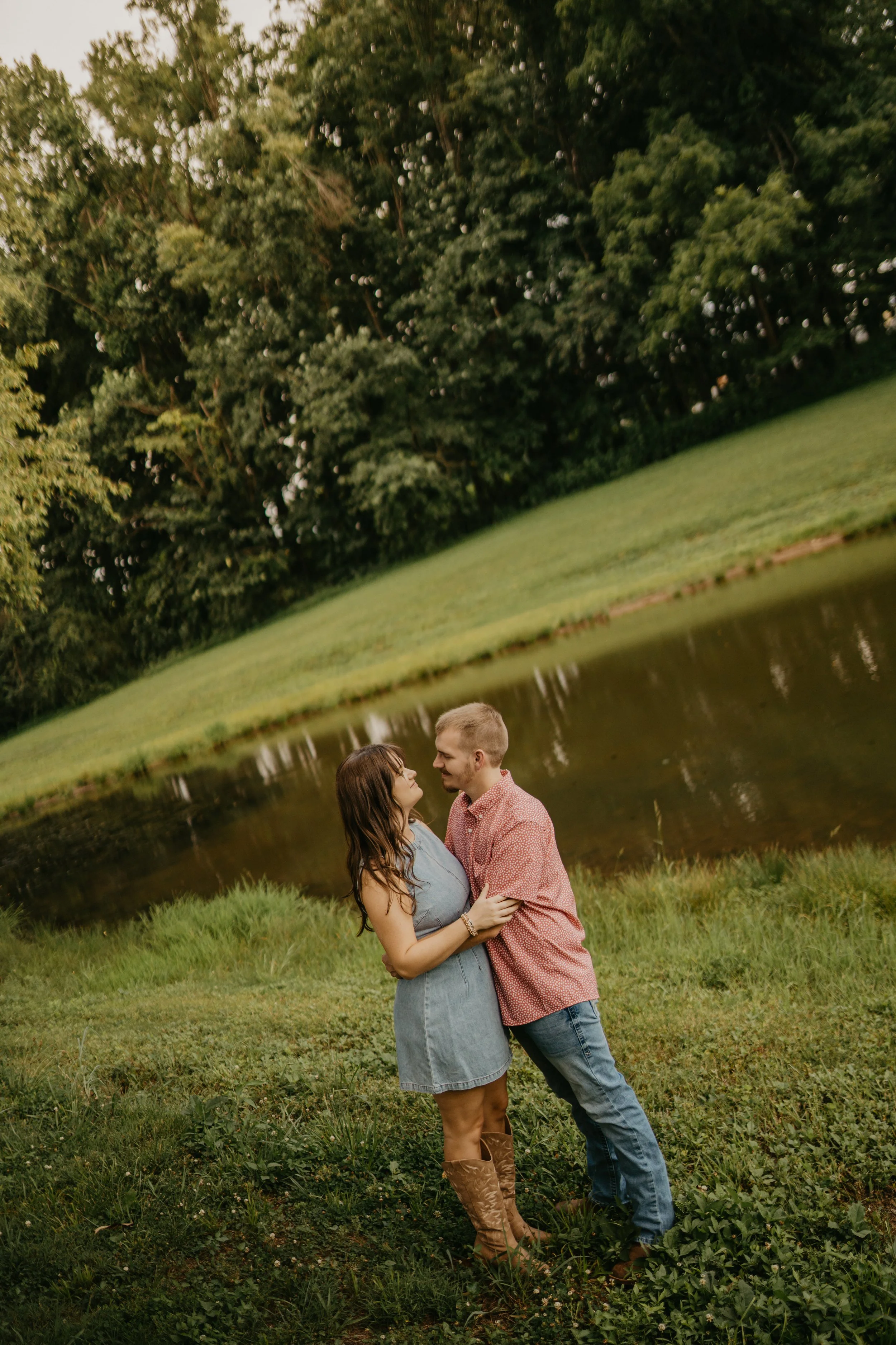 A couple standing close together outdoors by a pond, embracing, smiling, with trees in the background, during daytime.