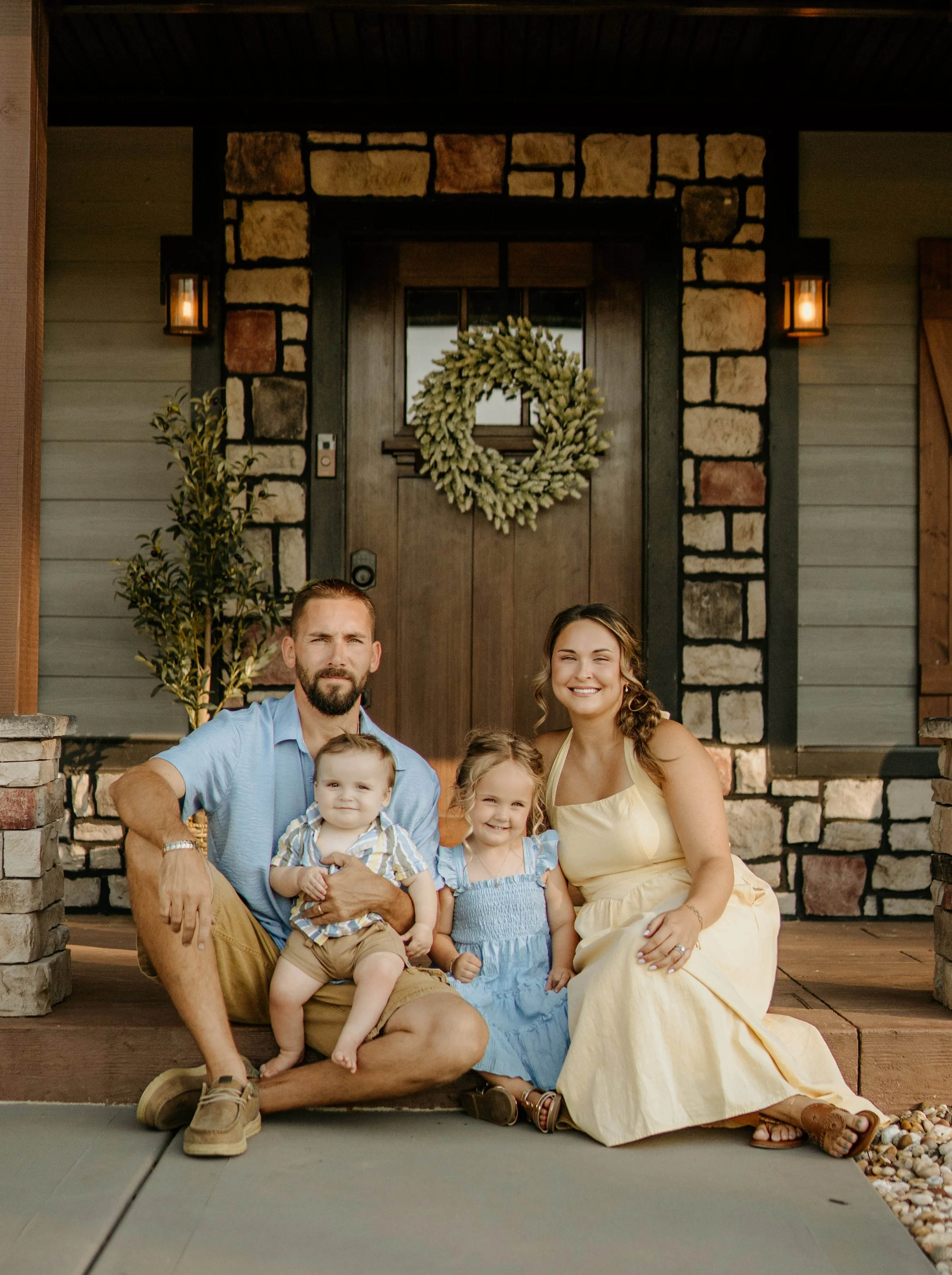 Family of five sitting on front porch of a house decorated with a wreath and lanterns, smiling at the camera.