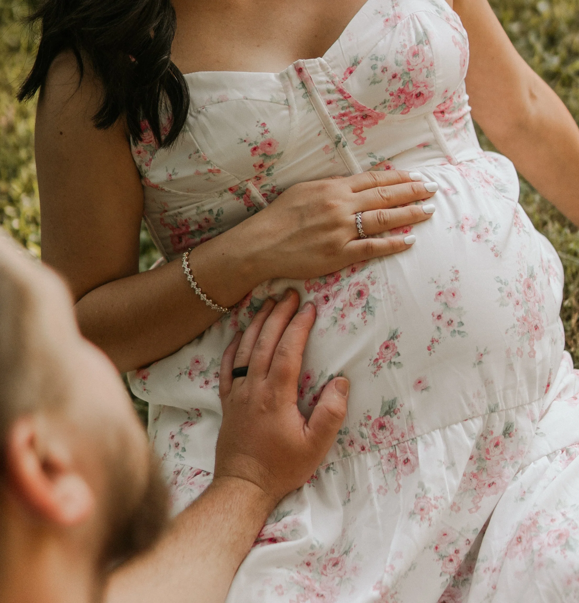 Close-up of a woman wearing a floral dress, with her hand resting on her pregnant belly, in an outdoor setting with greenery.