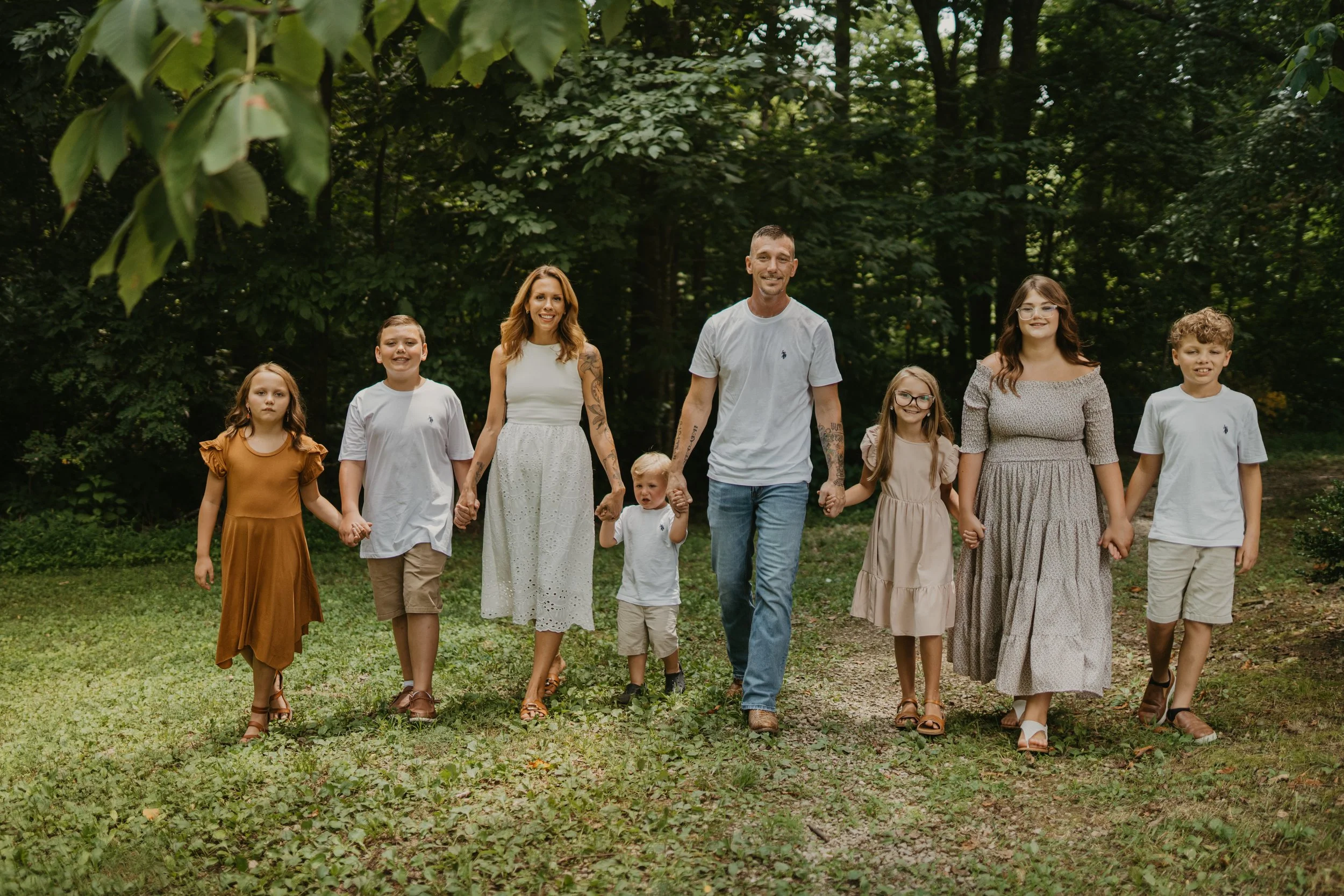 A family of eight people, including two adults and six children, walking hand in hand through a park surrounded by trees and greenery.