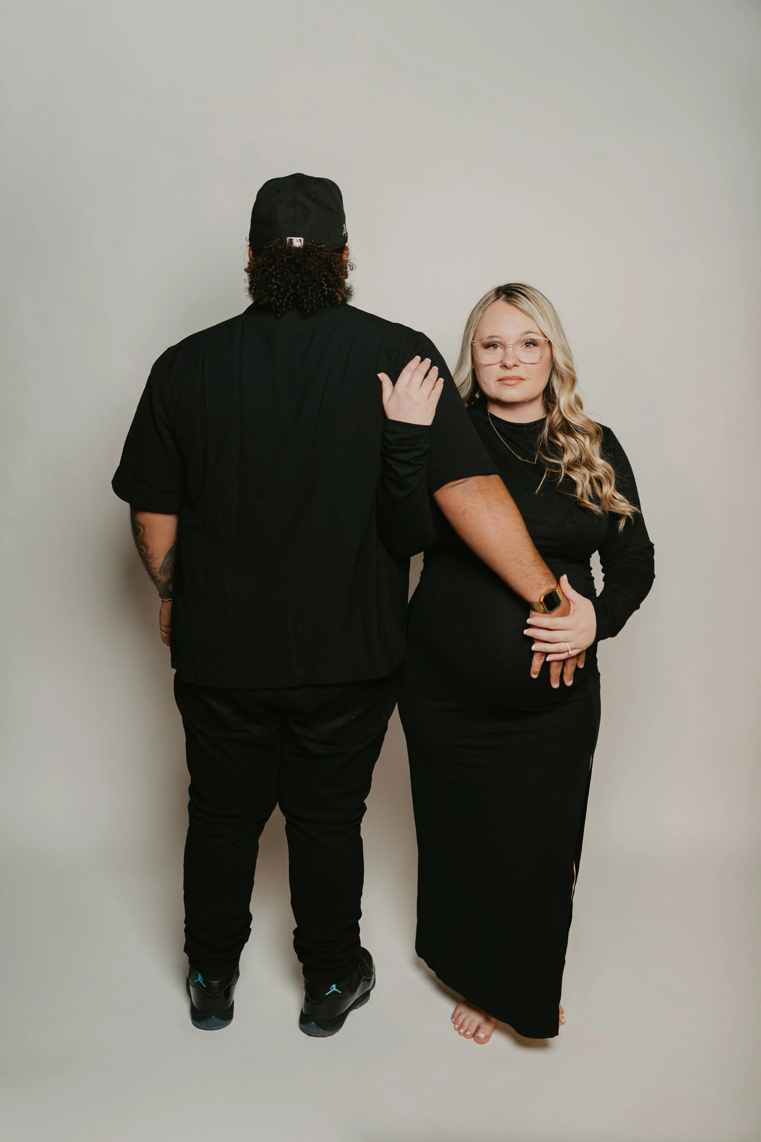 A man and a woman standing against a white background, facing away from each other. The man has curly hair, wears a black cap, black jacket, black pants, and sneakers. The woman has long blonde hair, glasses, and is dressed in a black dress, barefoot