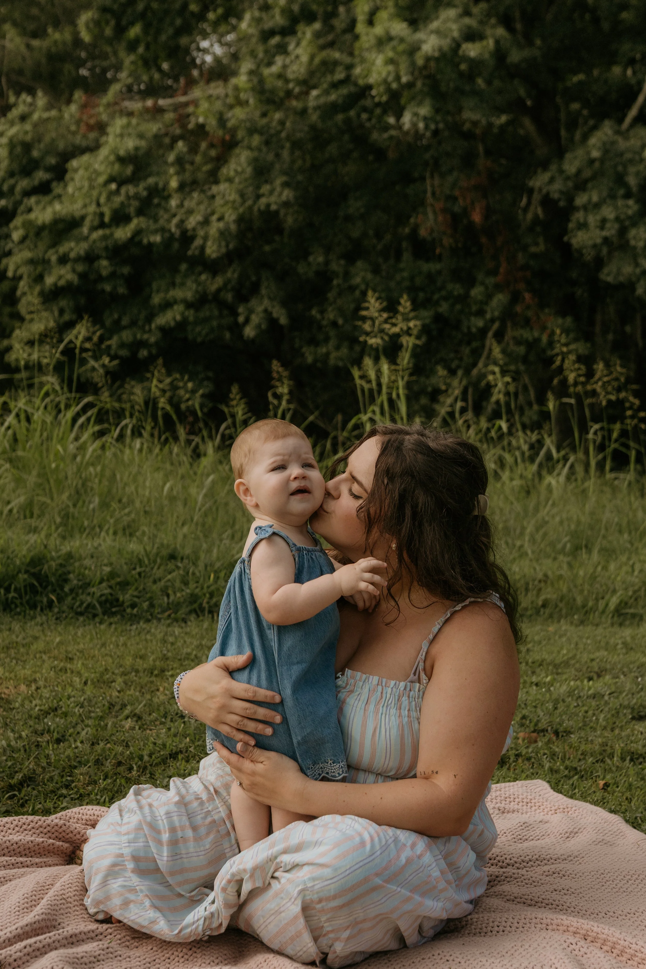 A woman with dark curly hair holding a toddler girl with blonde hair, both sitting on a pink blanket outdoors. The woman is kissing the girl, who looks slightly upset. Tall grass and trees are in the background.