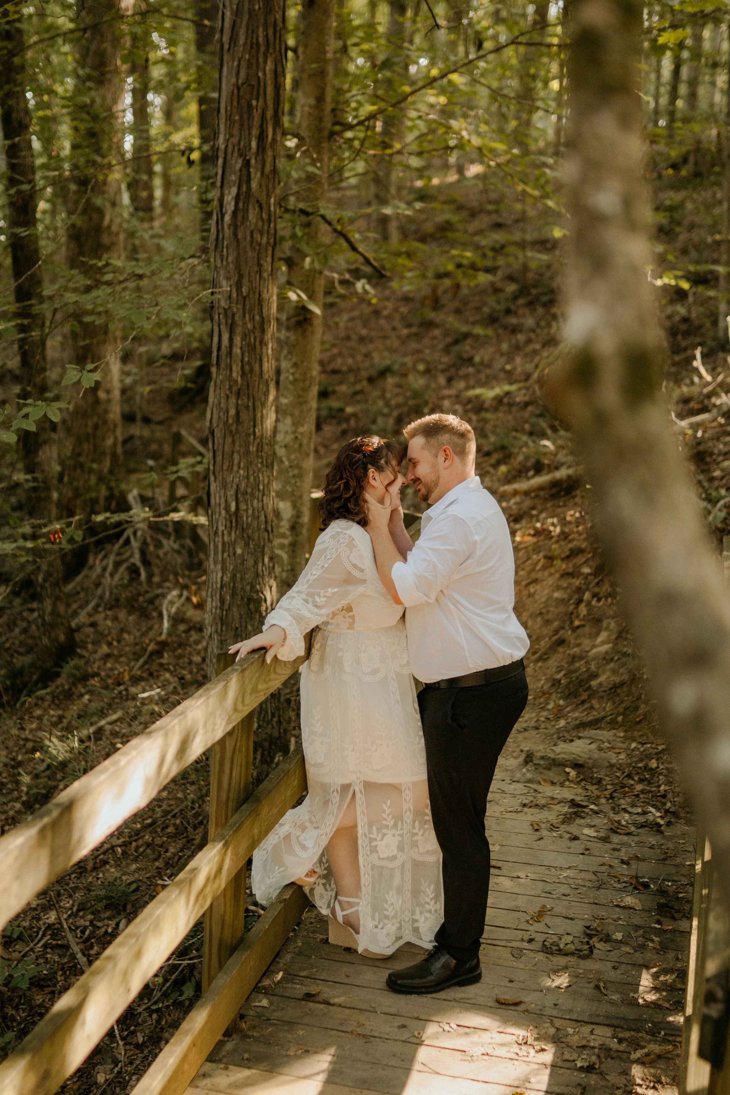 A couple stands close together on a wooden forest trail, touching foreheads and smiling, surrounded by trees and natural woodland scenery.