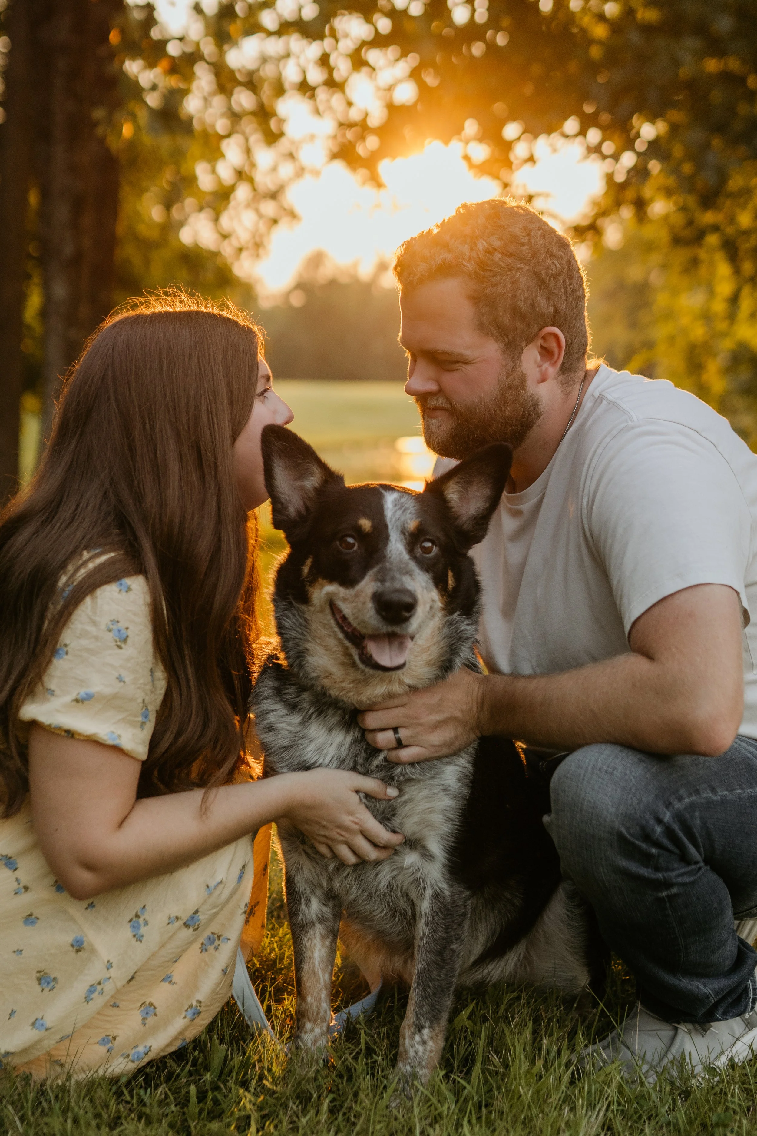 A couple and their dog enjoying sunset outdoors in a park, with trees and water in the background.