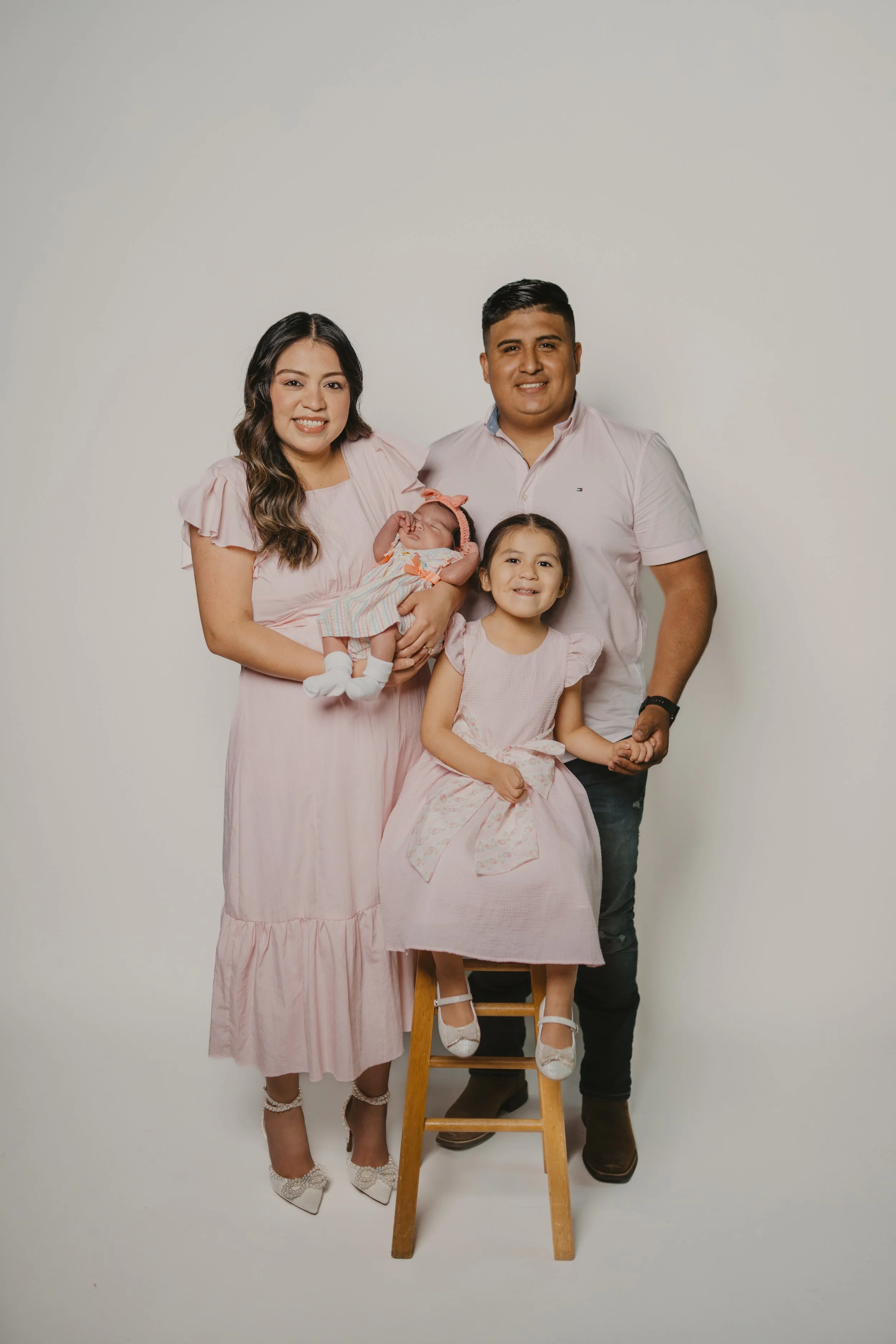 Family of four, including a woman holding a newborn, a man, and a young girl, standing in front of a plain white background. The woman is dressed in a pink dress and high heels, the man in a light pink shirt and dark pants, and the girl in a light pi