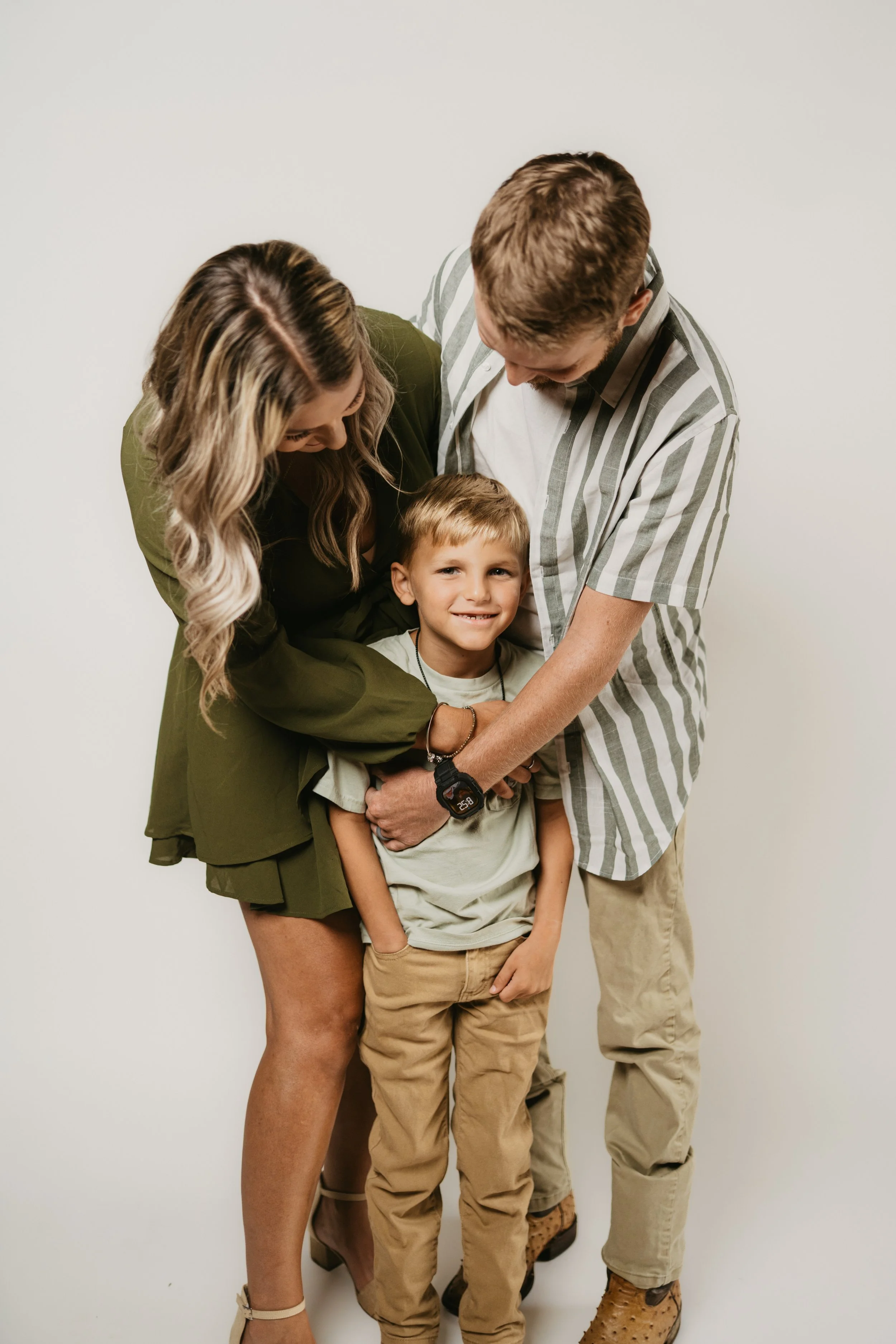 A smiling young boy being embraced by his parents against a plain white background.