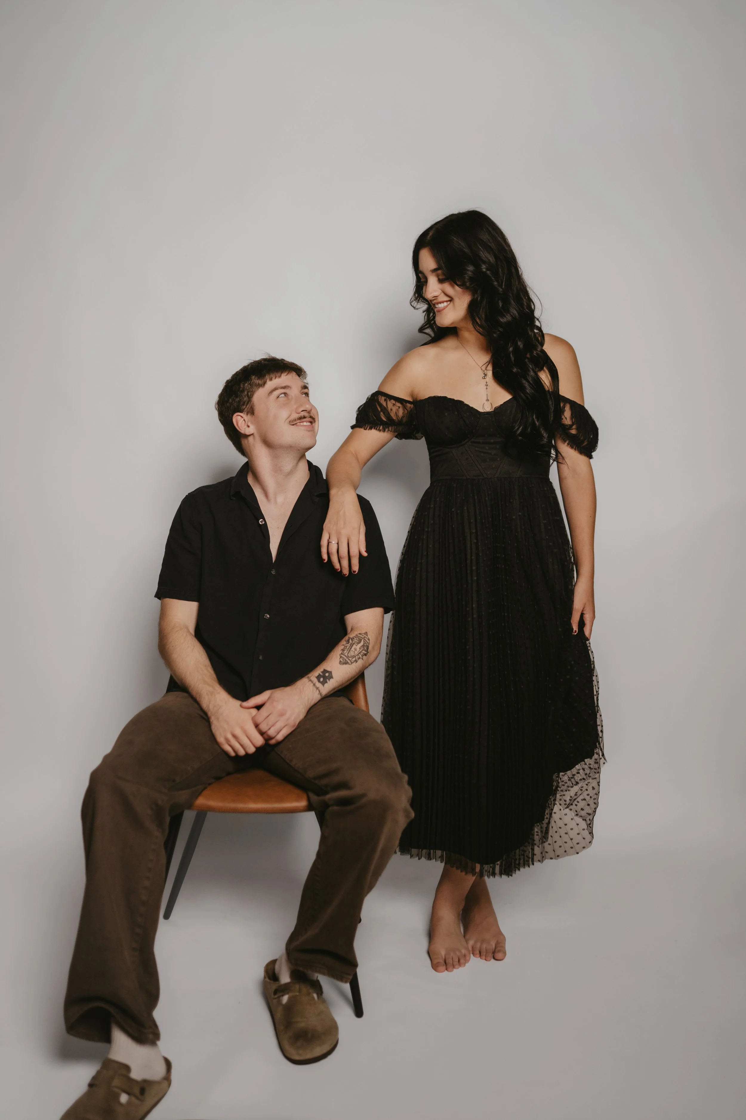A man sitting on a chair looking up at a woman standing beside him, both smiling. The woman is dressed in a black dress and the man is wearing a black shirt and brown pants. The background is plain white.