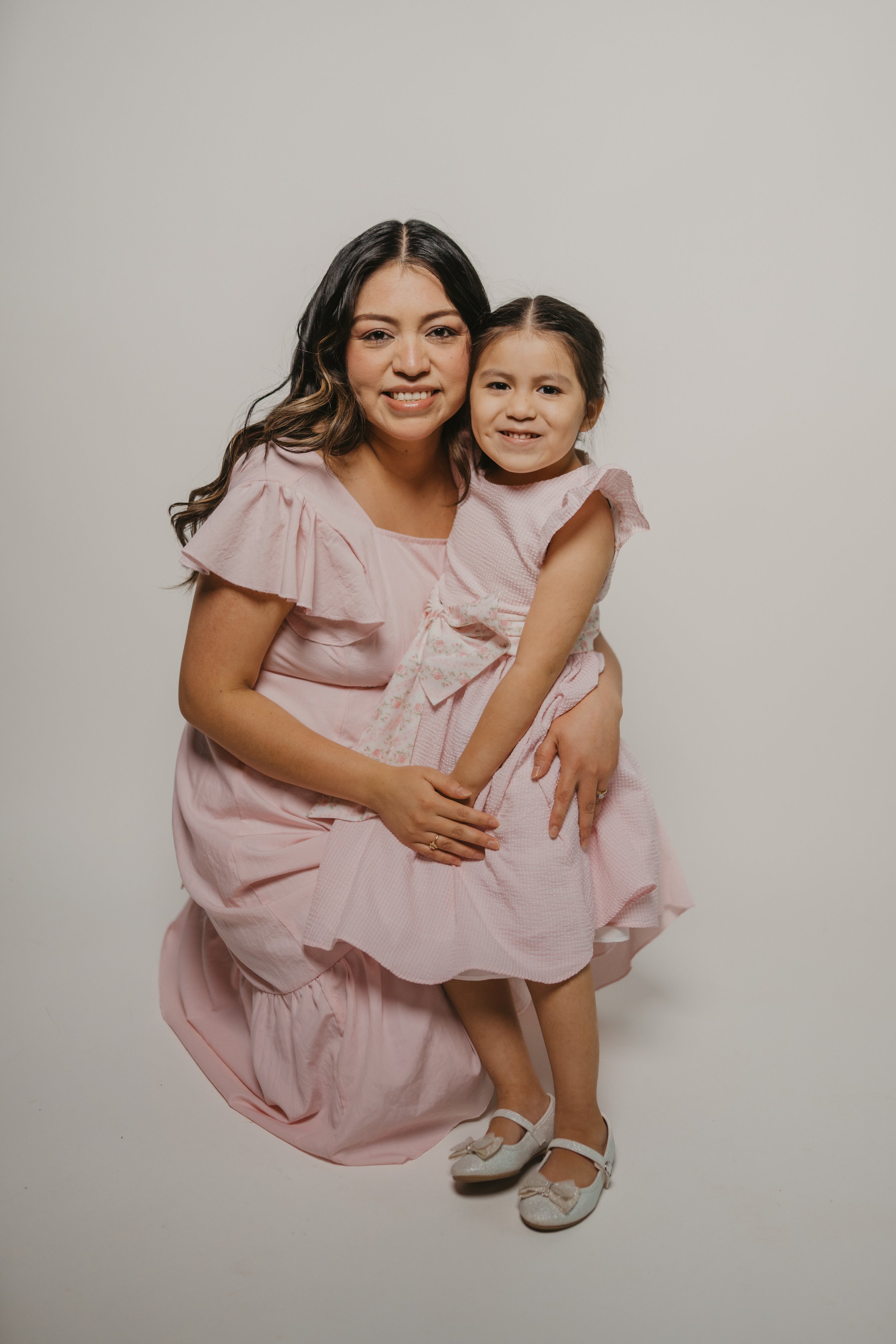 A woman with long dark hair and a young girl with braided hair, both wearing pink dresses, smiling and sitting against a plain white background.