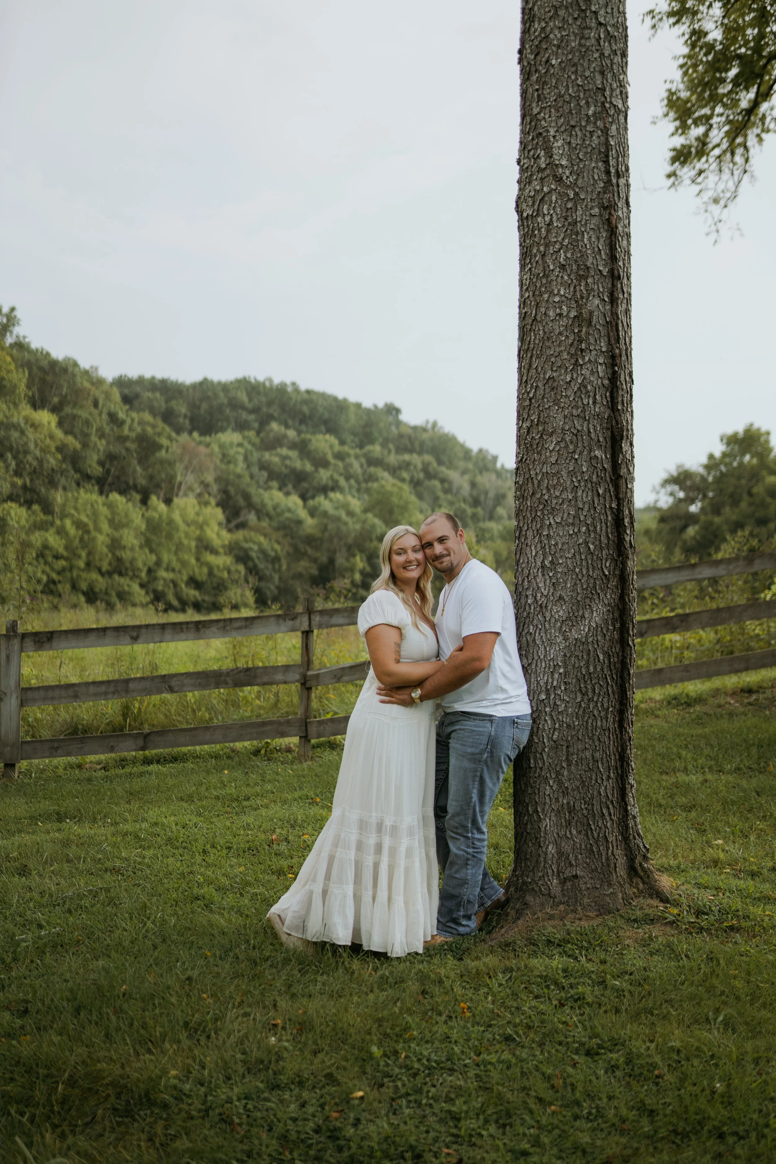 A couple stands close together outdoors near a large tree, smiling at the camera, with a wooden fence and green hills in the background.