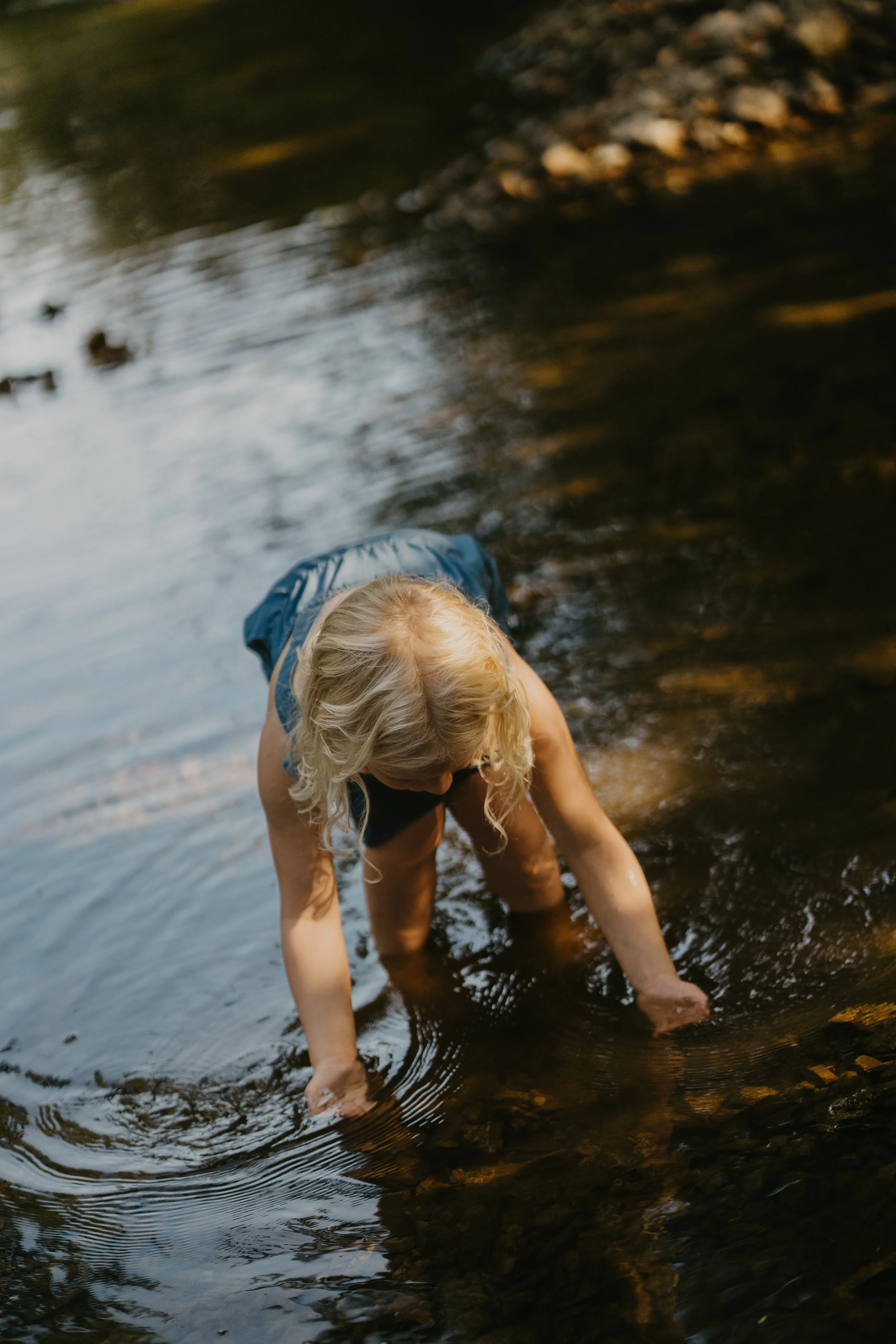 A person with blonde hair wearing a sleeveless shirt and shorts, bent over and touching a shallow body of water with their hands, in a natural outdoor setting.