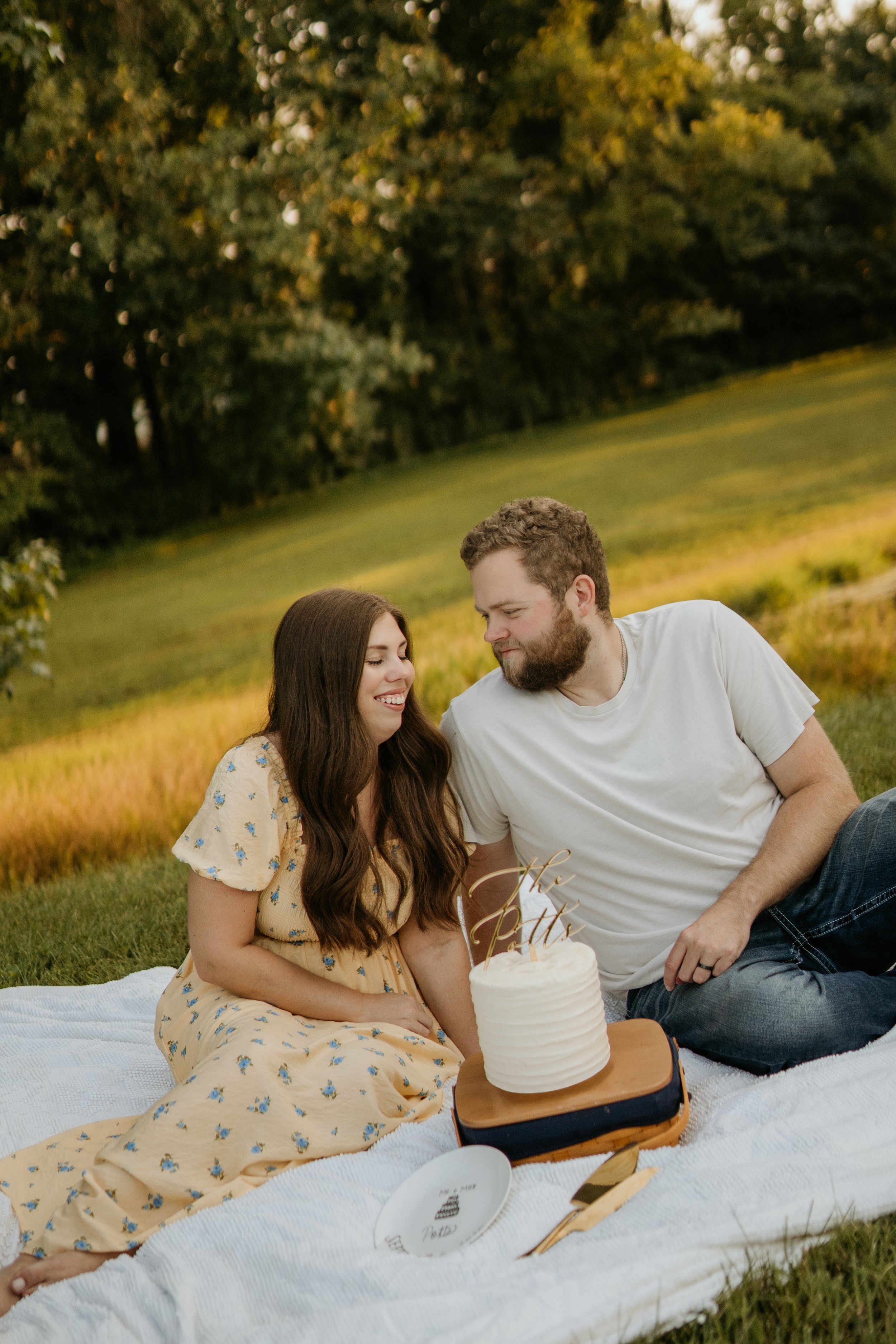 A couple sitting on a white blanket on the grass outdoors, celebrating a special occasion with a small white cake and a 'The Pola's' cake topper, surrounded by a scenic background of trees and grassy fields.