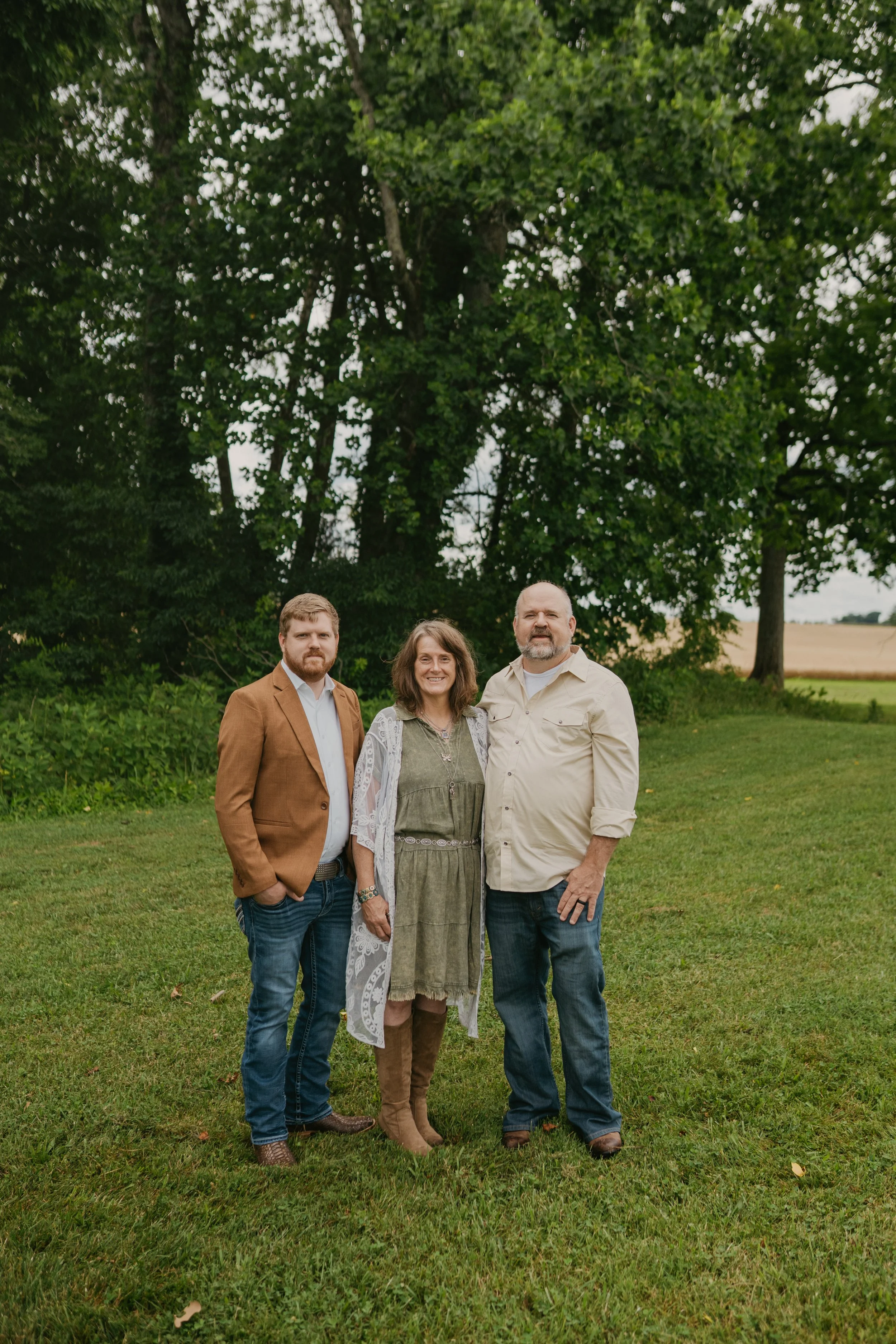 A group of three adults, two men and one woman, standing outdoors on grass with trees in the background, during daytime. They are smiling and dressed in casual and semi-formal clothing.