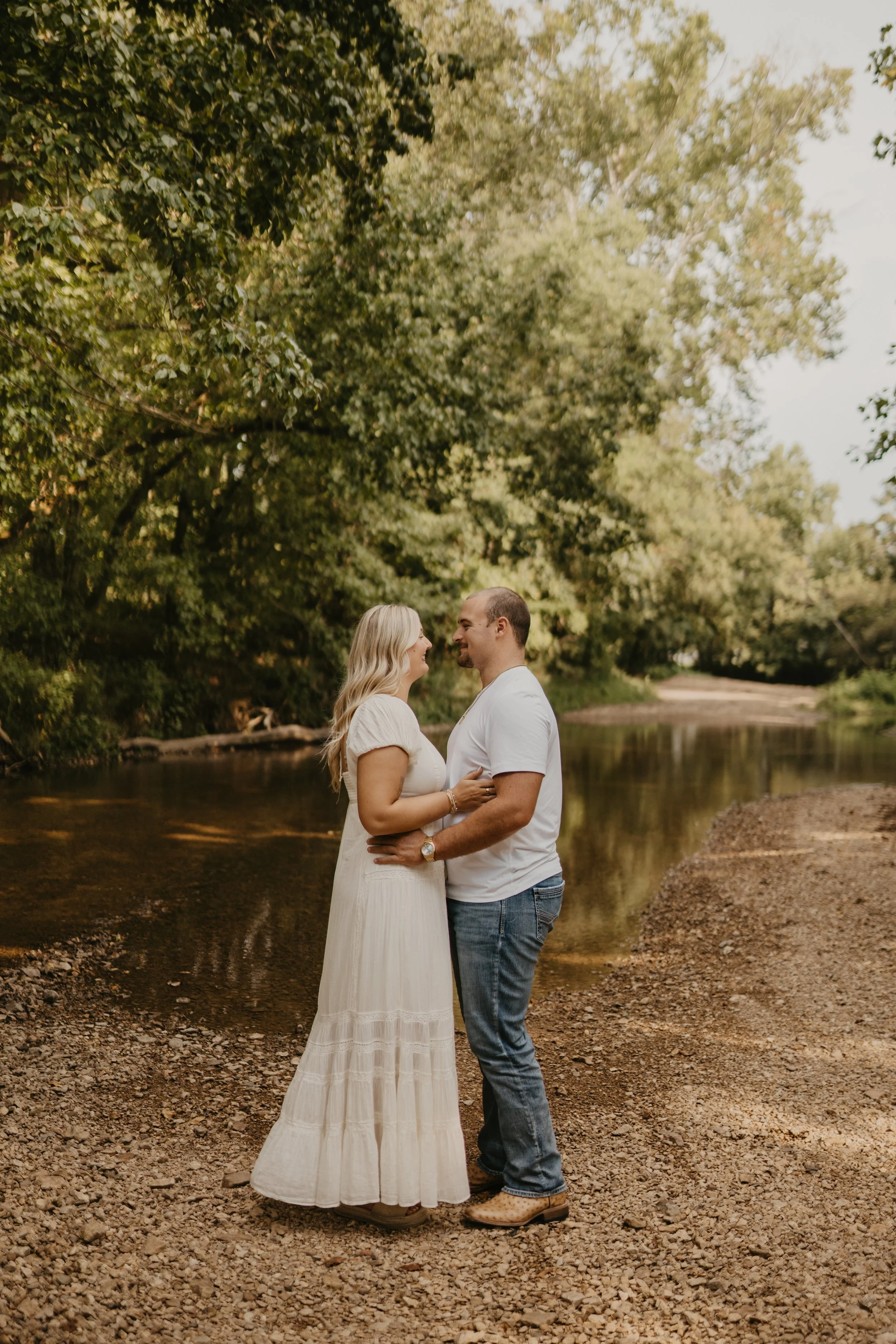 A couple stands close together outdoors near a creek, smiling and looking into each other's eyes, surrounded by green trees and rocky ground.