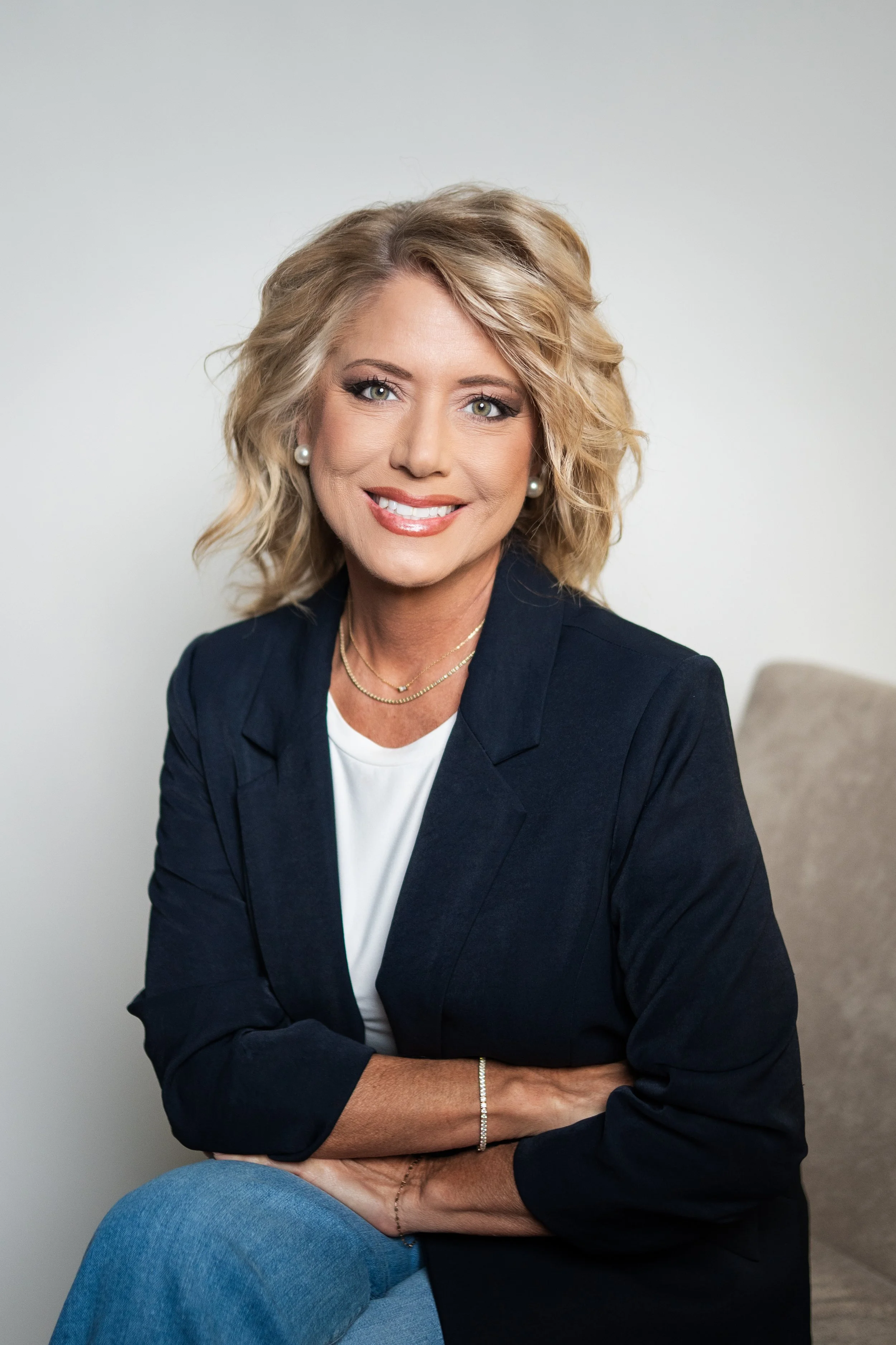 Portrait of a woman with blonde, wavy hair, wearing a black blazer over a white top, accessorized with pearl earrings and layered necklaces, sitting against a plain light background.