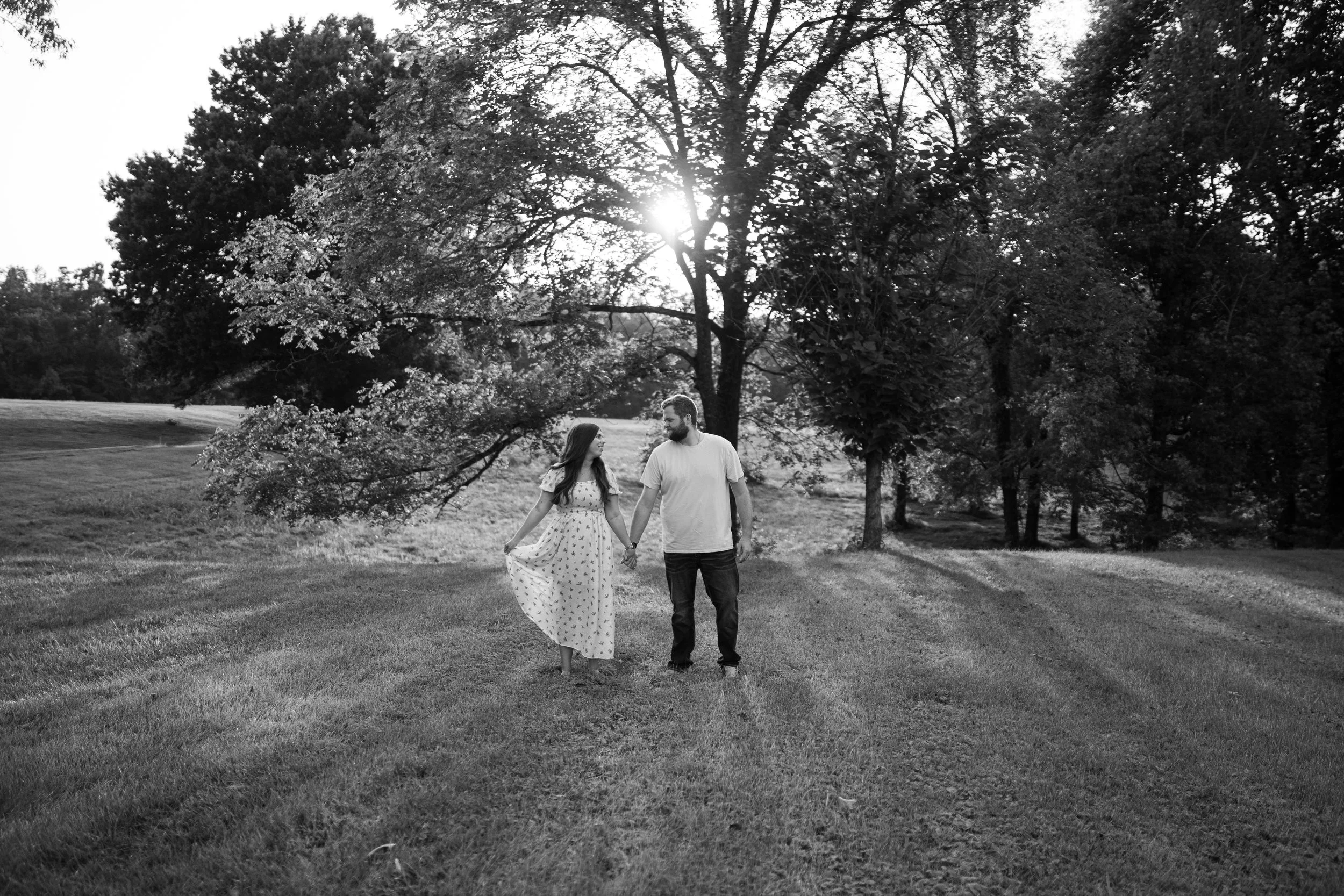 Black and white photo of a man and woman holding hands and walking in a park with trees, grass, and sunlight shining through the trees.