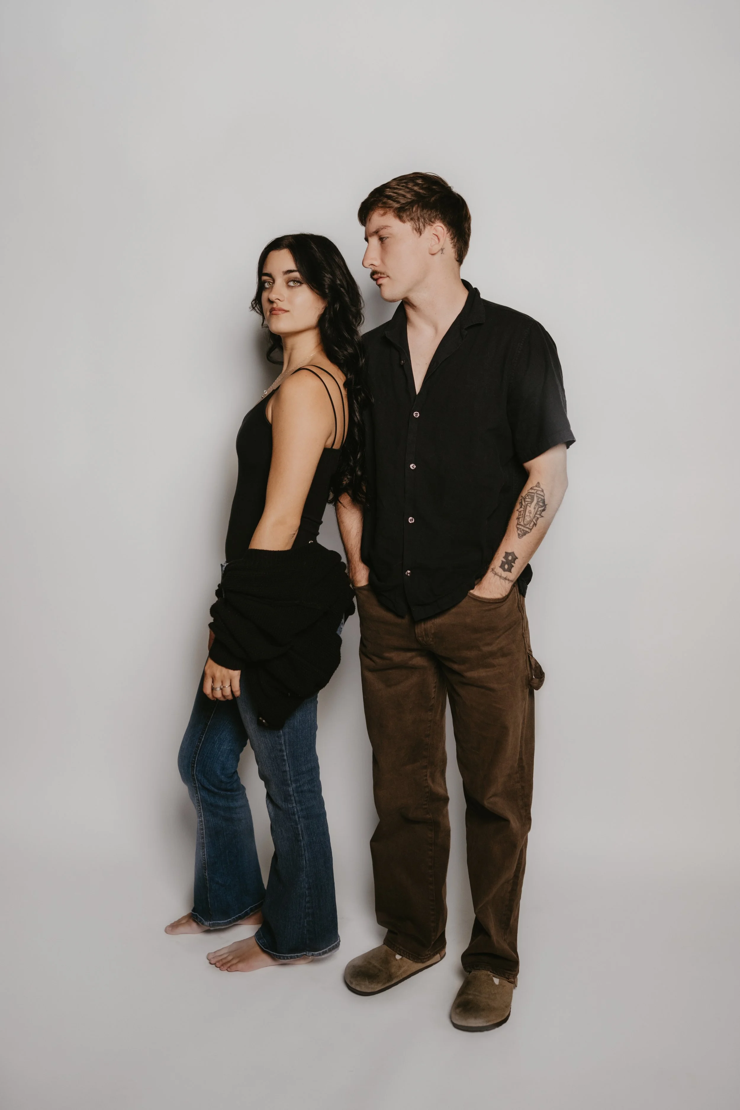 A man and woman standing side by side against a plain white background. The woman has long black hair, is wearing a black sleeveless top and blue jeans. The man has short brown hair, facial hair, and is wearing a black button-up shirt with brown pant