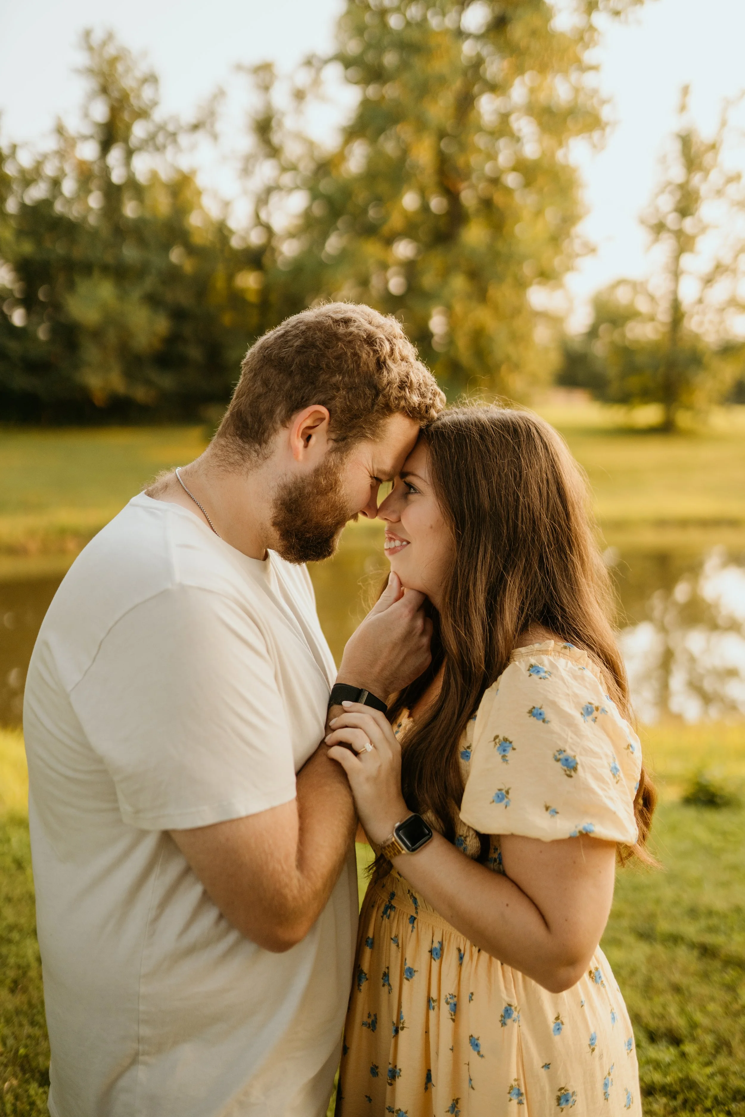 A couple in a park with trees in the background, touching foreheads and smiling at each other.