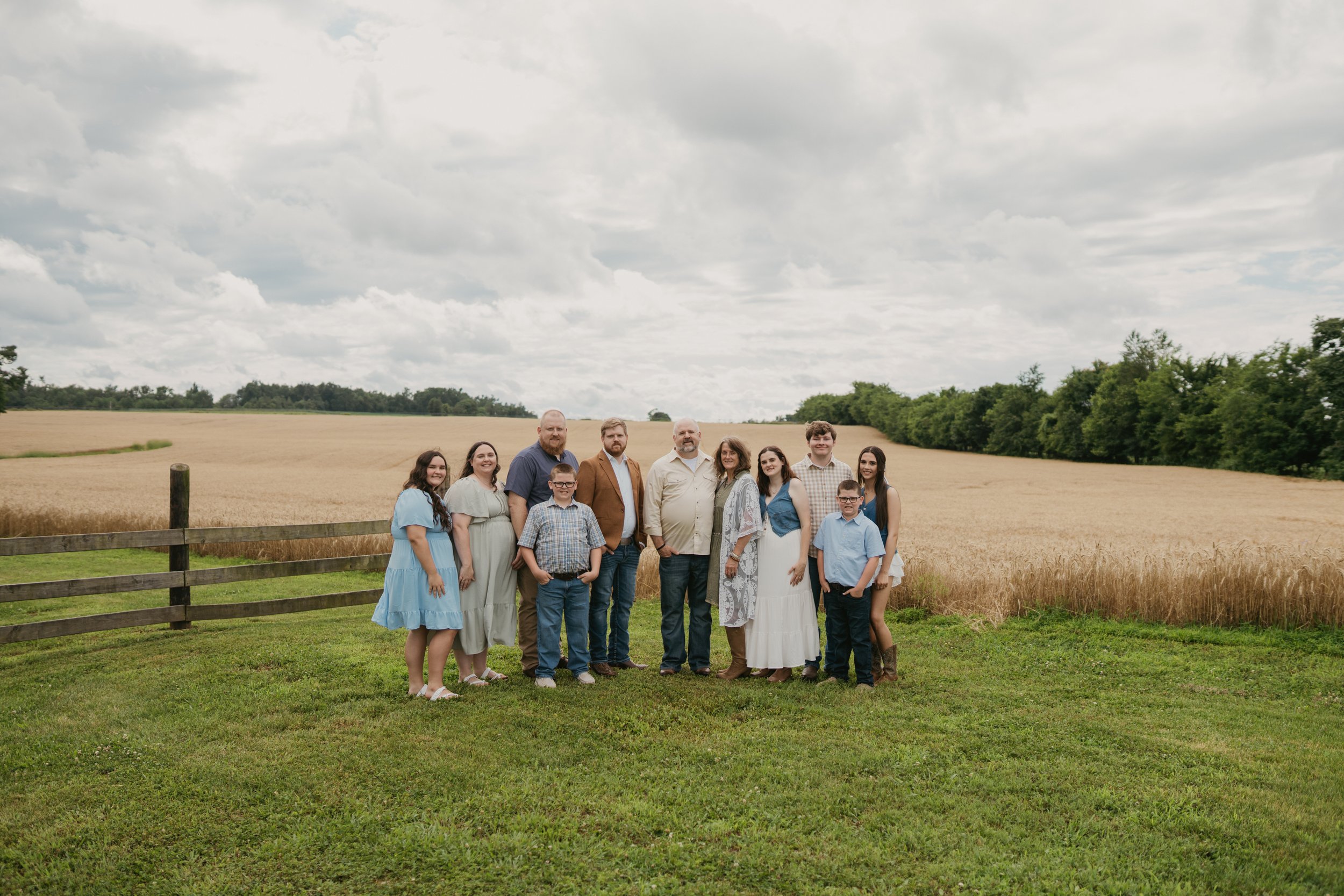 Group of people standing on grassy area near wheat field under cloudy sky, posing for a photo.